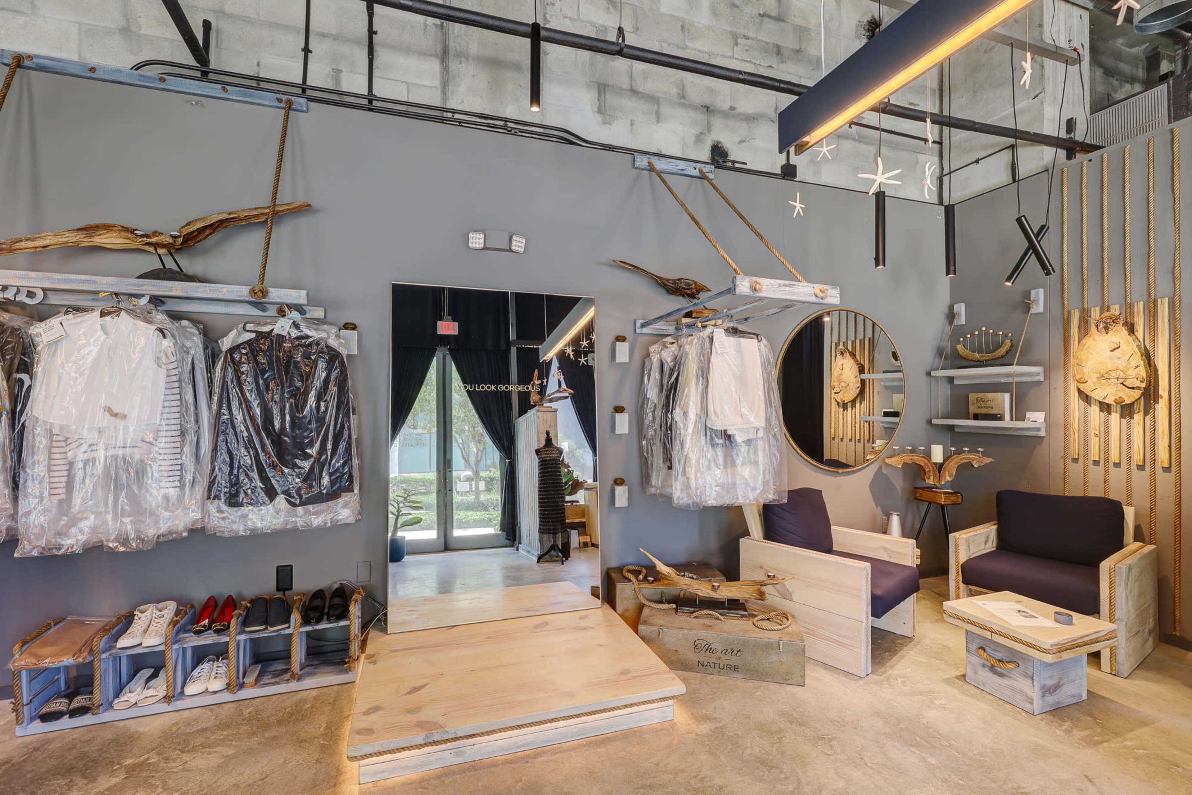 The image shows a modern clothing store interior featuring hanging garments, a full-length mirror, minimalist seating, and wooden display fixtures against a gray wall.