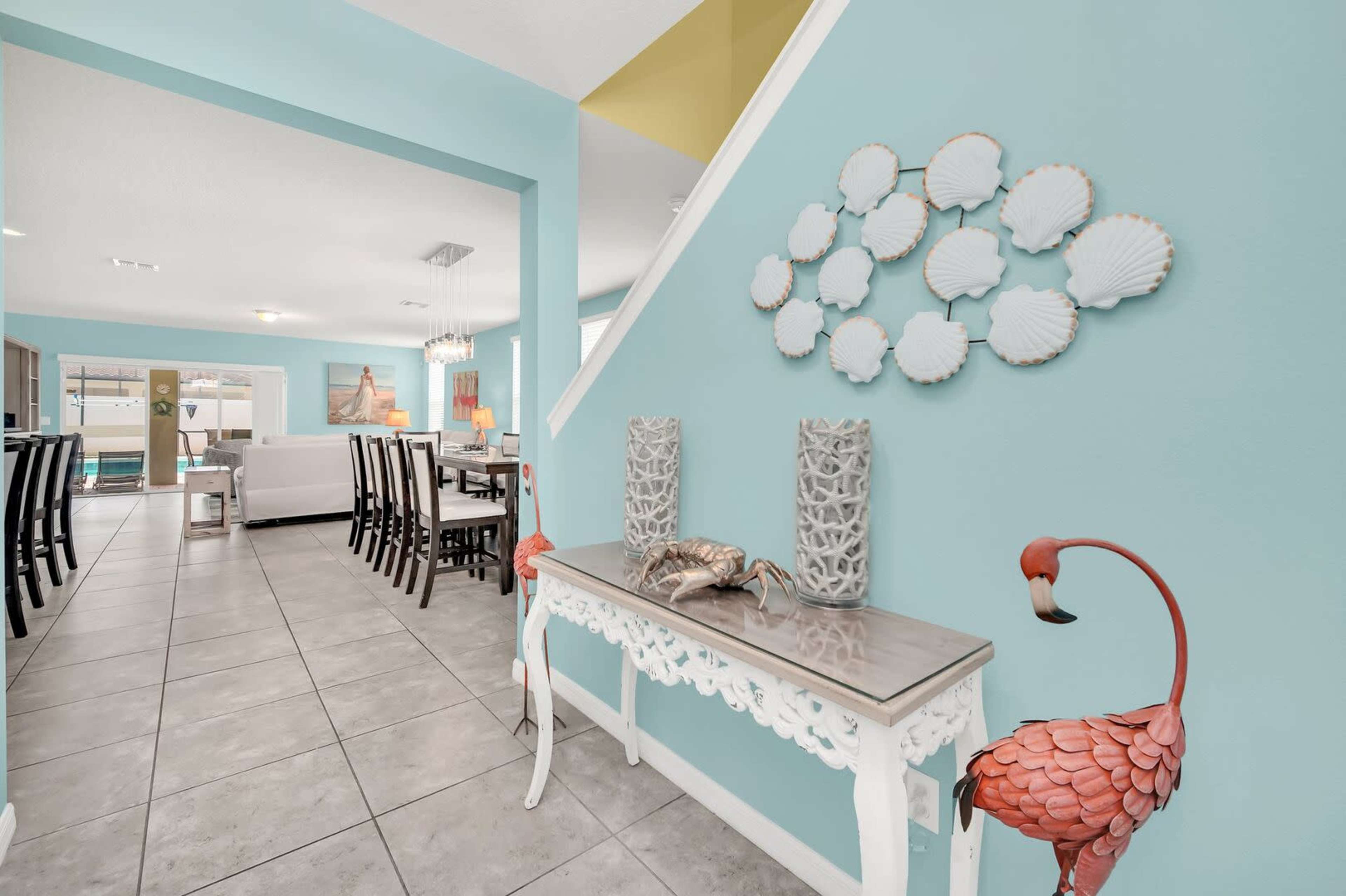 A brightly colored room with a blue wall, a decorative shell display, a console table with two flamingo statues, and a view of a living area in the background.