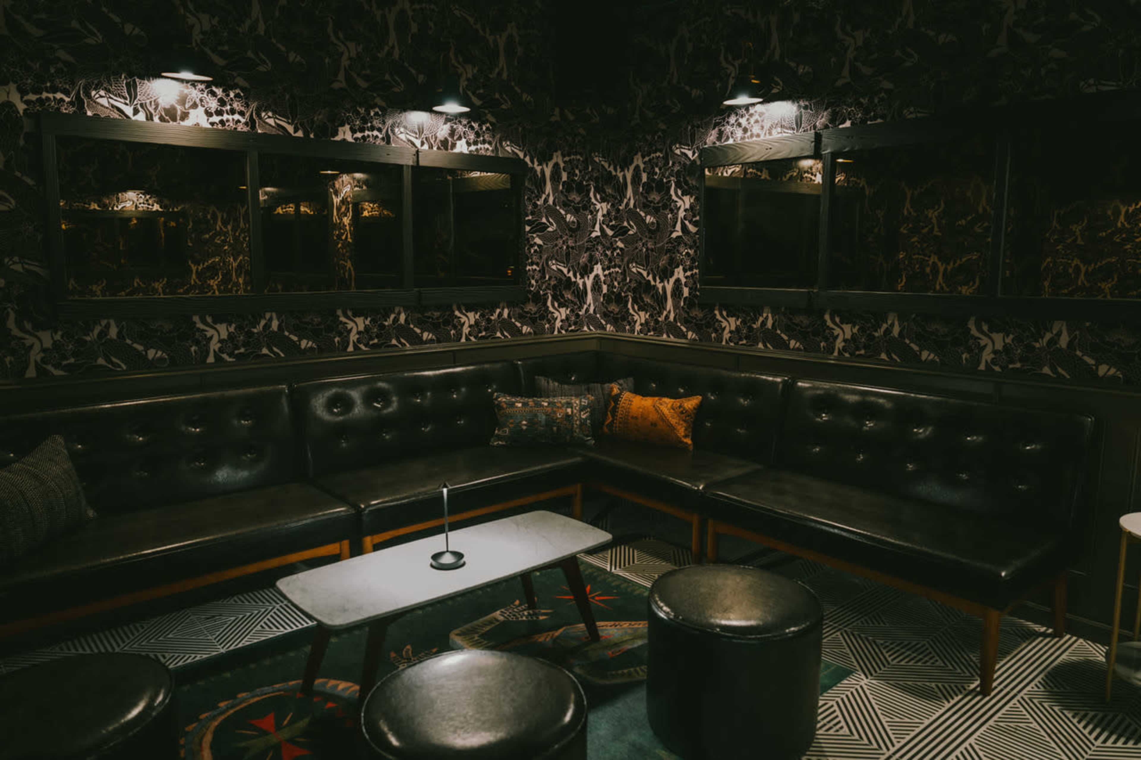 The image depicts a dimly lit lounge area featuring a black leather sectional sofa, round stools, and patterned wallpaper with mirrors along one wall.