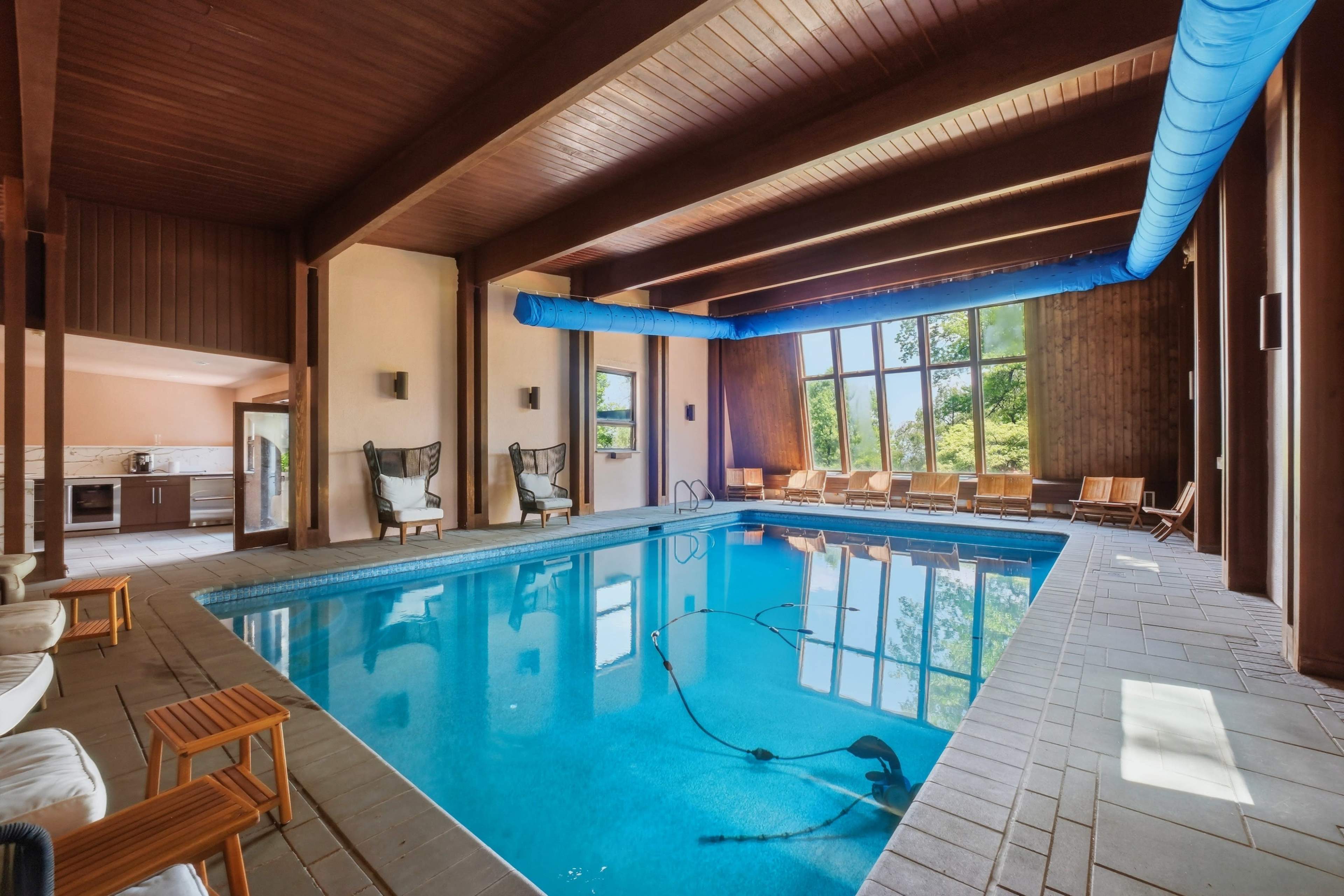 The image shows an indoor swimming pool area with large windows, wooden beams, and lounge chairs arranged along the edges.
