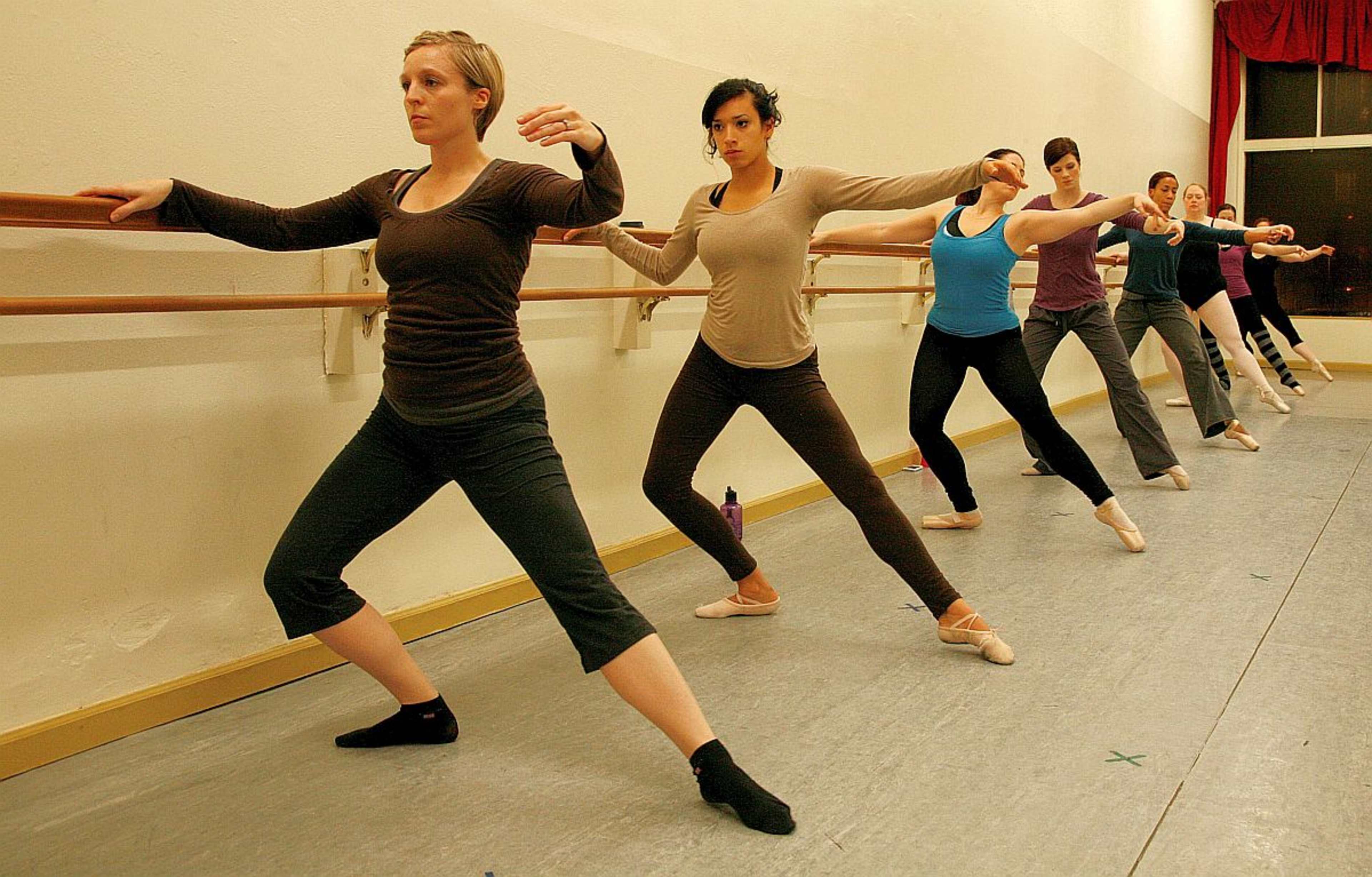 A group of dancers performs a ballet exercise at a studio, with each dancer facing the same direction along the ballet barre.