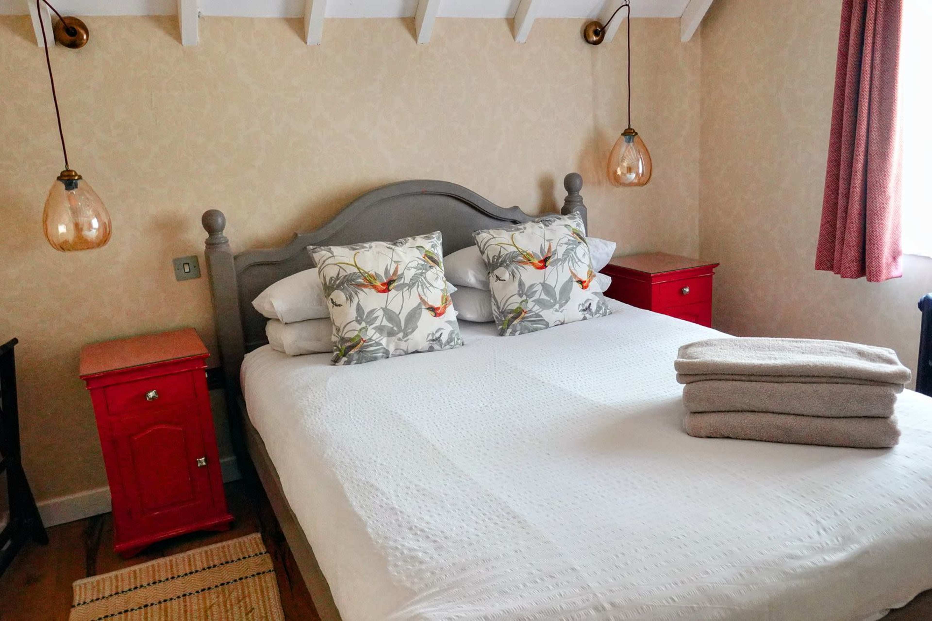 The image shows a neatly arranged bedroom with a double bed, decorative pillows, and red nightstands on either side.