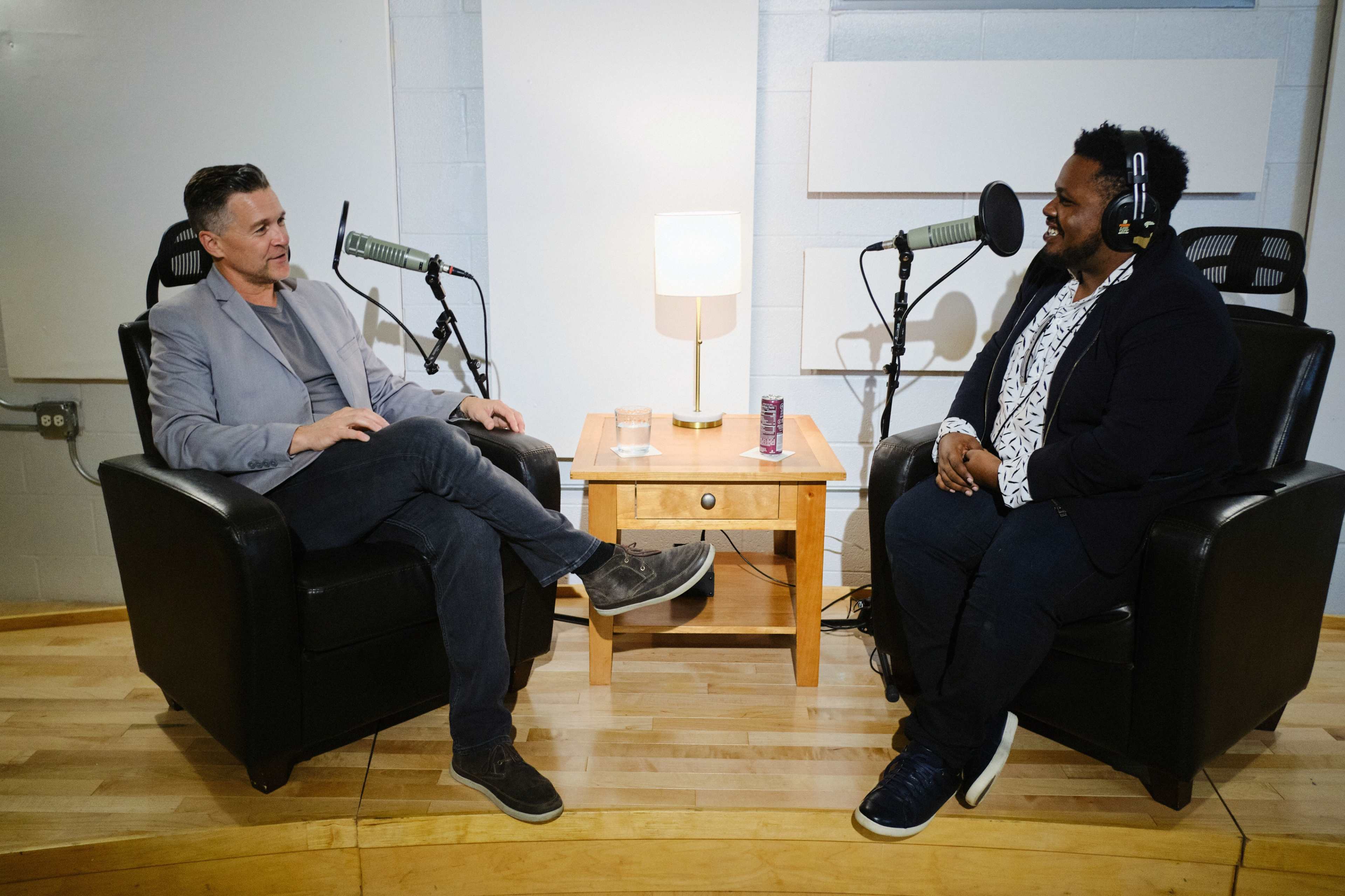 Two men are seated in black leather armchairs across from each other, engaging in conversation with microphones positioned between them and a small table with a lamp in the center.