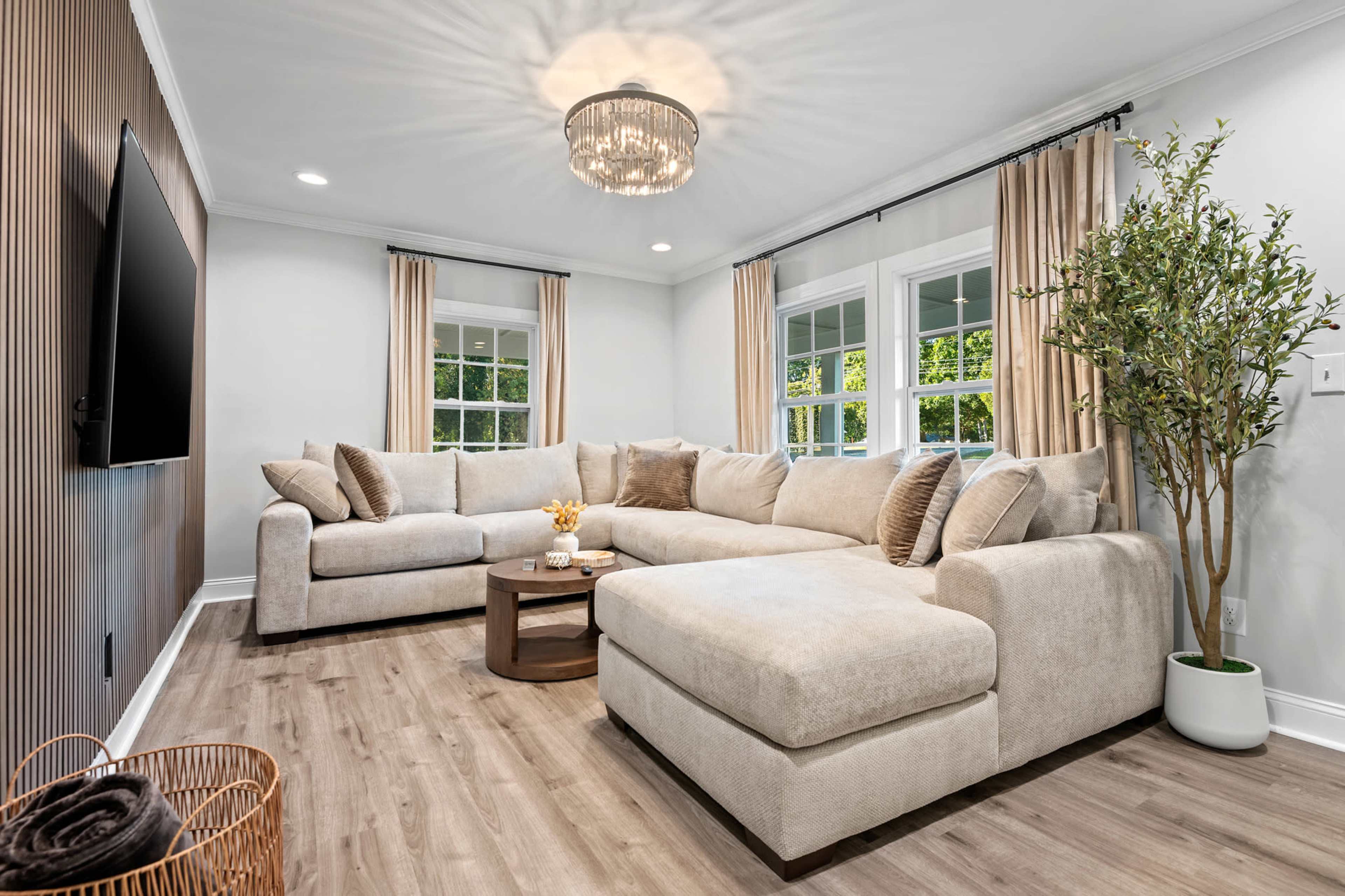 A spacious living room features a large, light-colored sectional sofa, a circular coffee table, and a decorative plant near the window, with a wall-mounted television opposite the seating area.