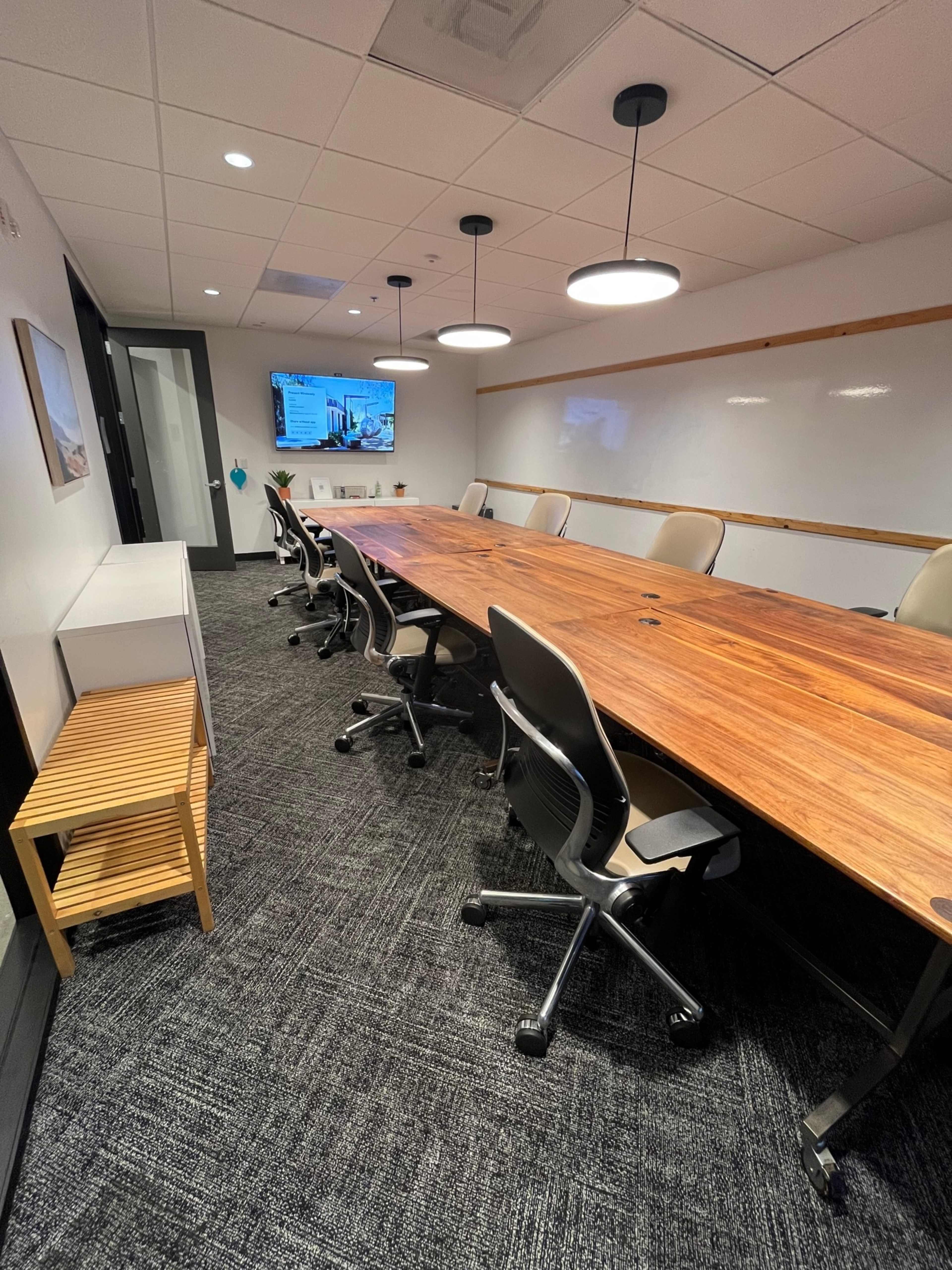 A conference room with a long wooden table, several ergonomic chairs, and a television mounted on the wall.