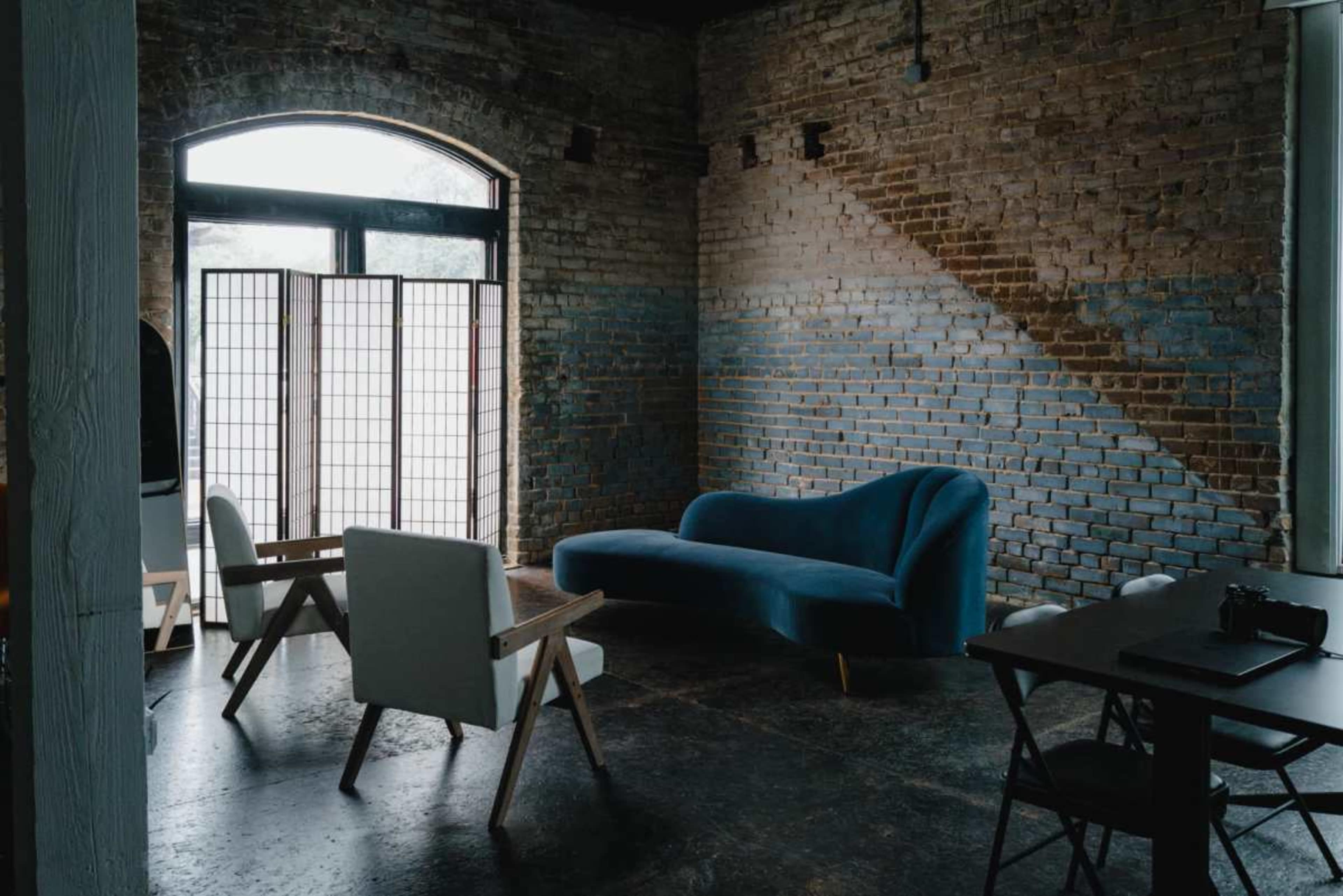 The image shows a minimalist room with a blue couch, two white chairs, and a folding room divider against a brick wall.