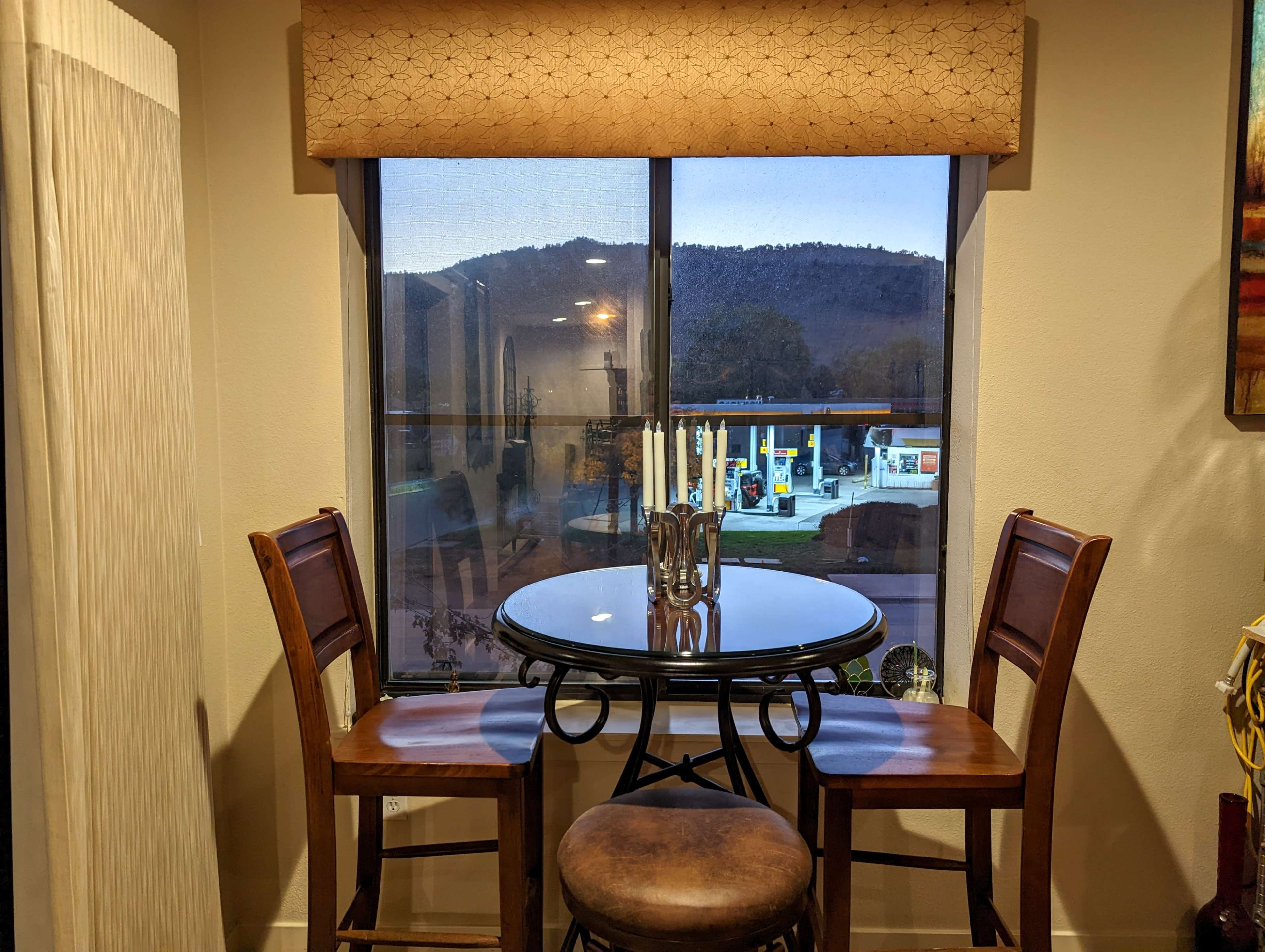 A small round table with two chairs is positioned near a window that overlooks a gas station and mountains in the background.
