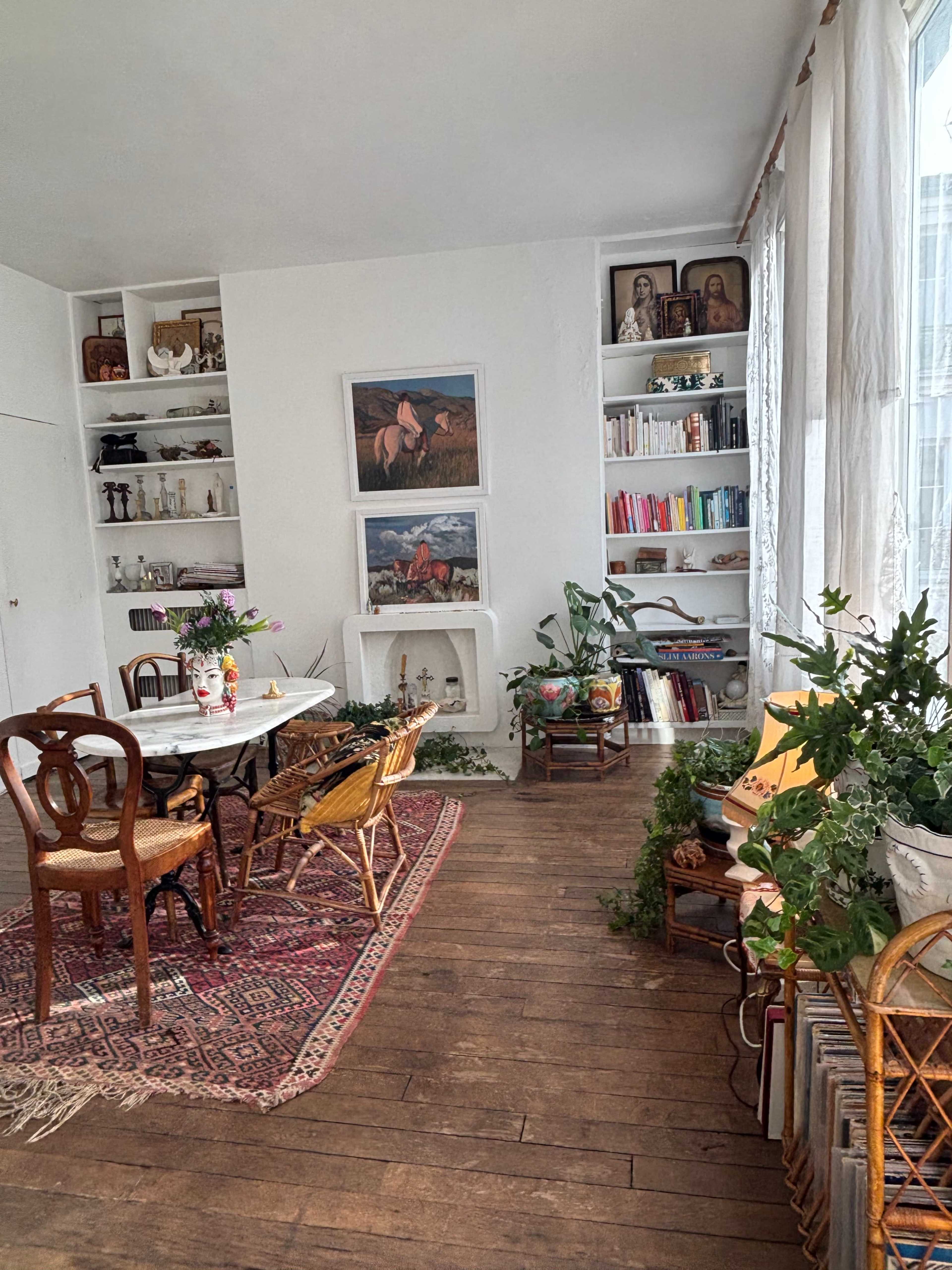 The image depicts a cozy room with a dining area featuring a table surrounded by chairs, bookshelves filled with books and decorative items, and plants scattered throughout.