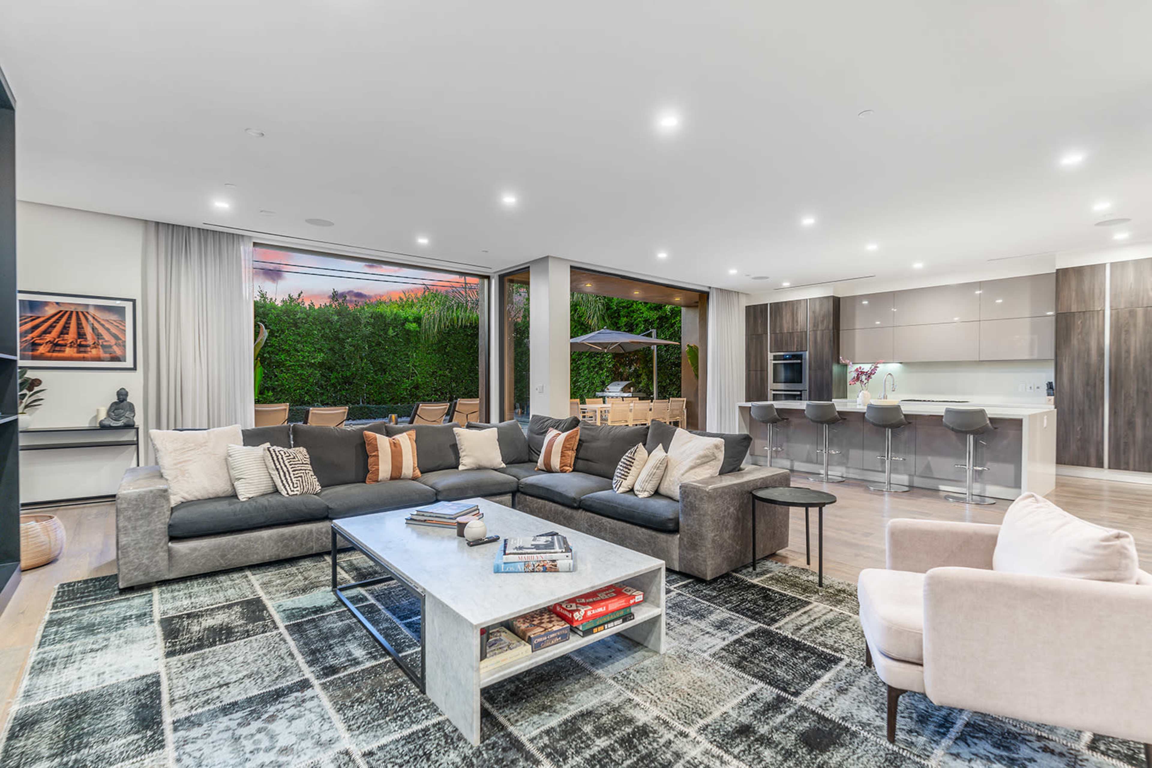The image shows a modern living room with a large gray sectional sofa, a coffee table with books, and a view of an outdoor patio through large sliding glass doors.