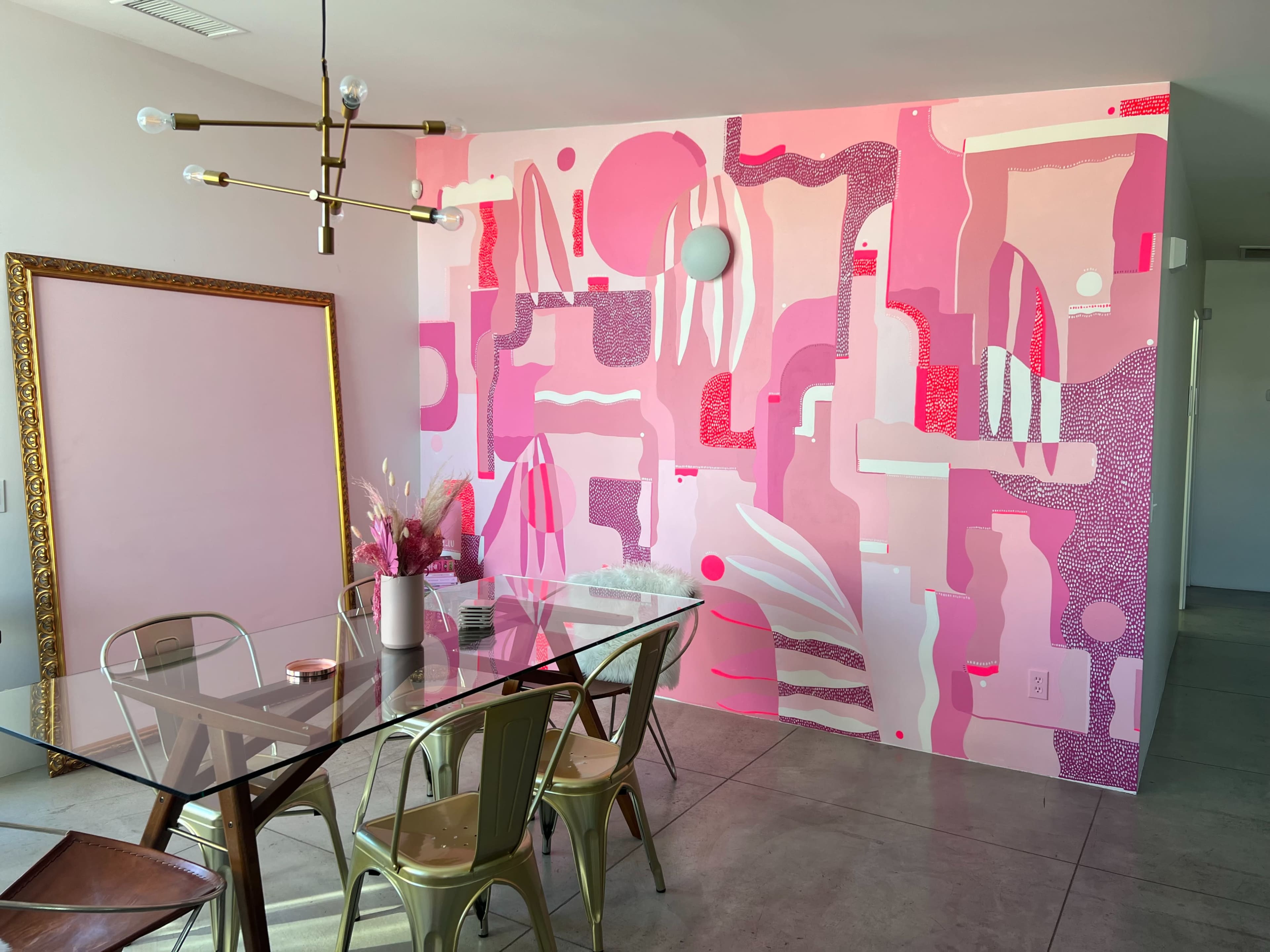 A dining area features a glass table surrounded by metal chairs, with a large pink abstract mural on the wall and a framed mirror nearby.