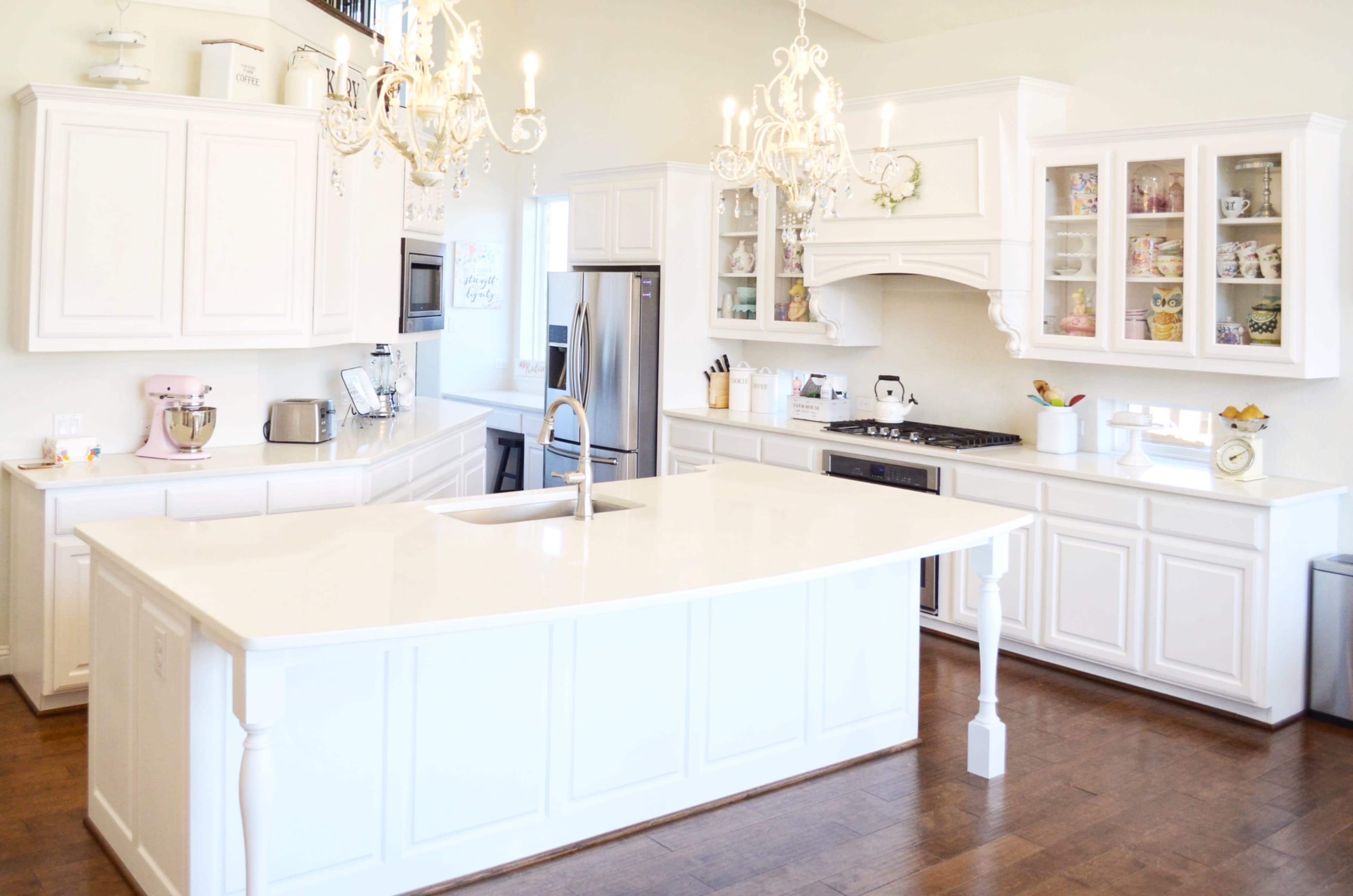 A spacious kitchen features white cabinetry, a large island with a sink, stainless steel appliances, and two chandeliers hanging from the ceiling.