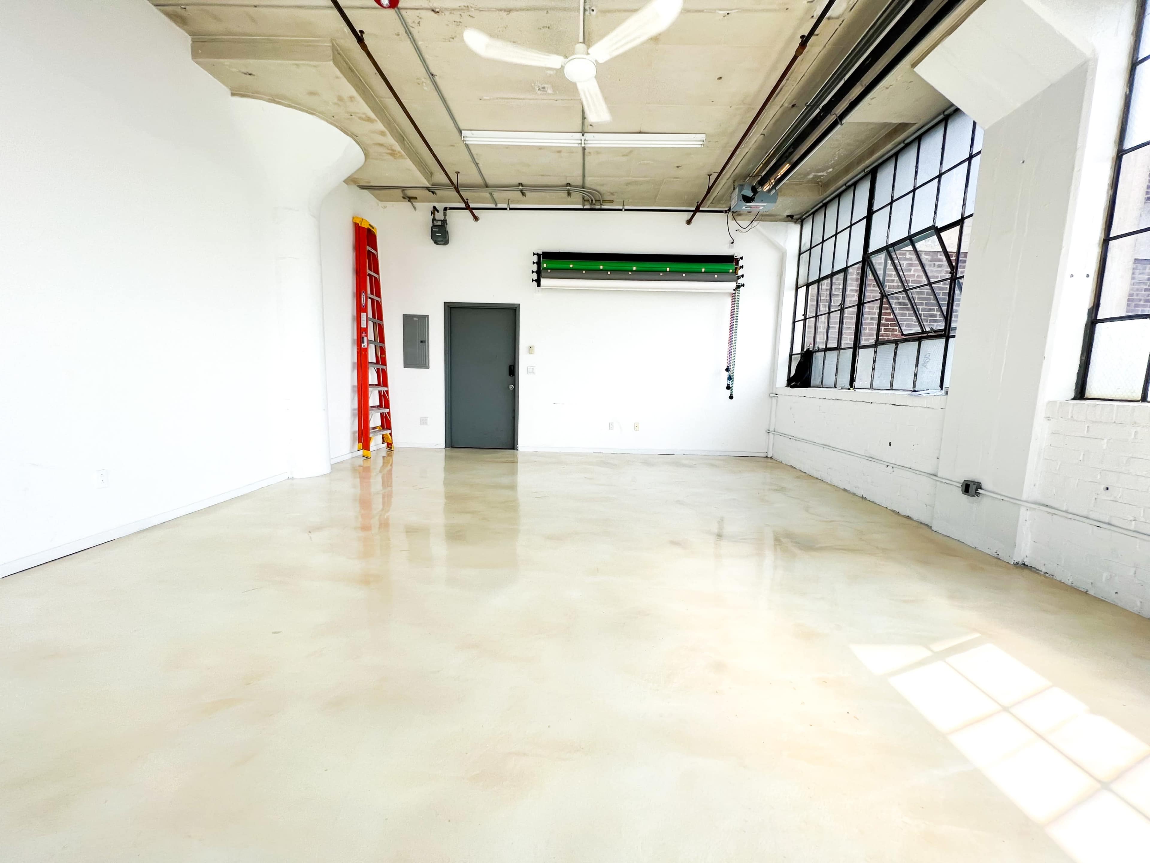 A spacious, empty room with concrete flooring, large windows, a small door, and a ladder against the wall.