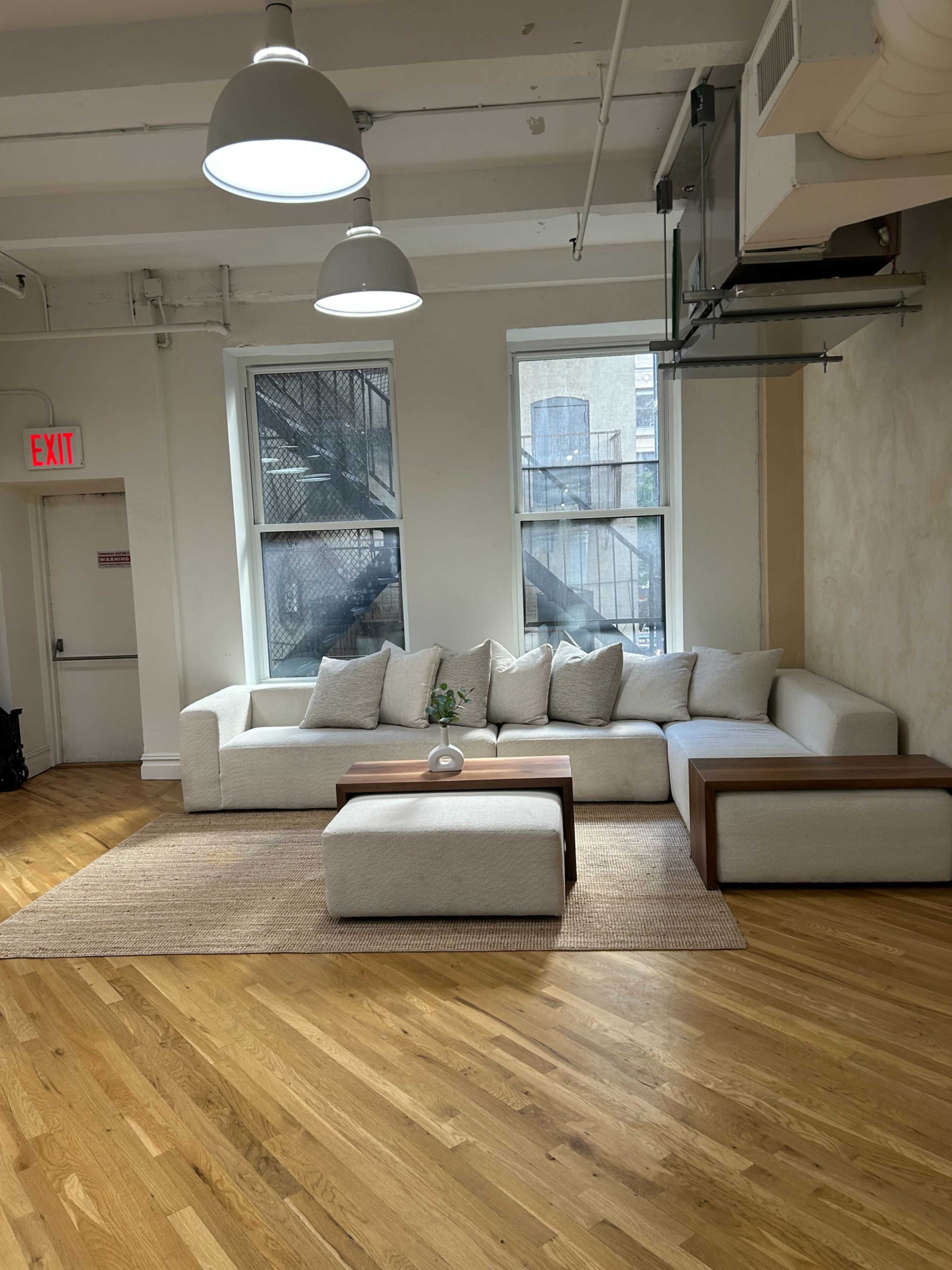 The image shows a modern, brightly lit living space featuring a large white sectional sofa with beige cushions, a wooden coffee table, and a woven area rug, positioned near two windows.