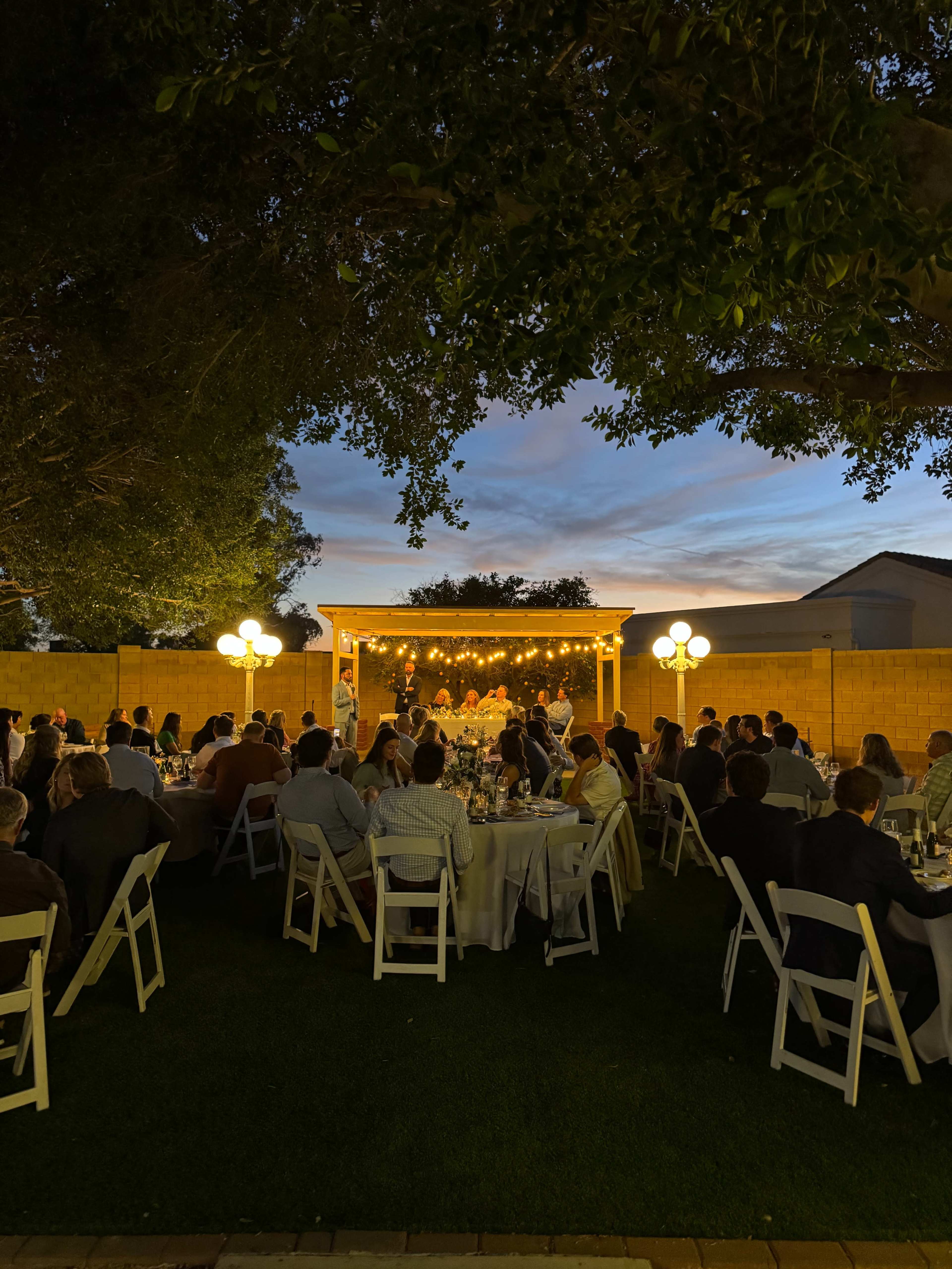 A backyard gathering features guests seated at round tables, with a stage illuminated by string lights in the background.