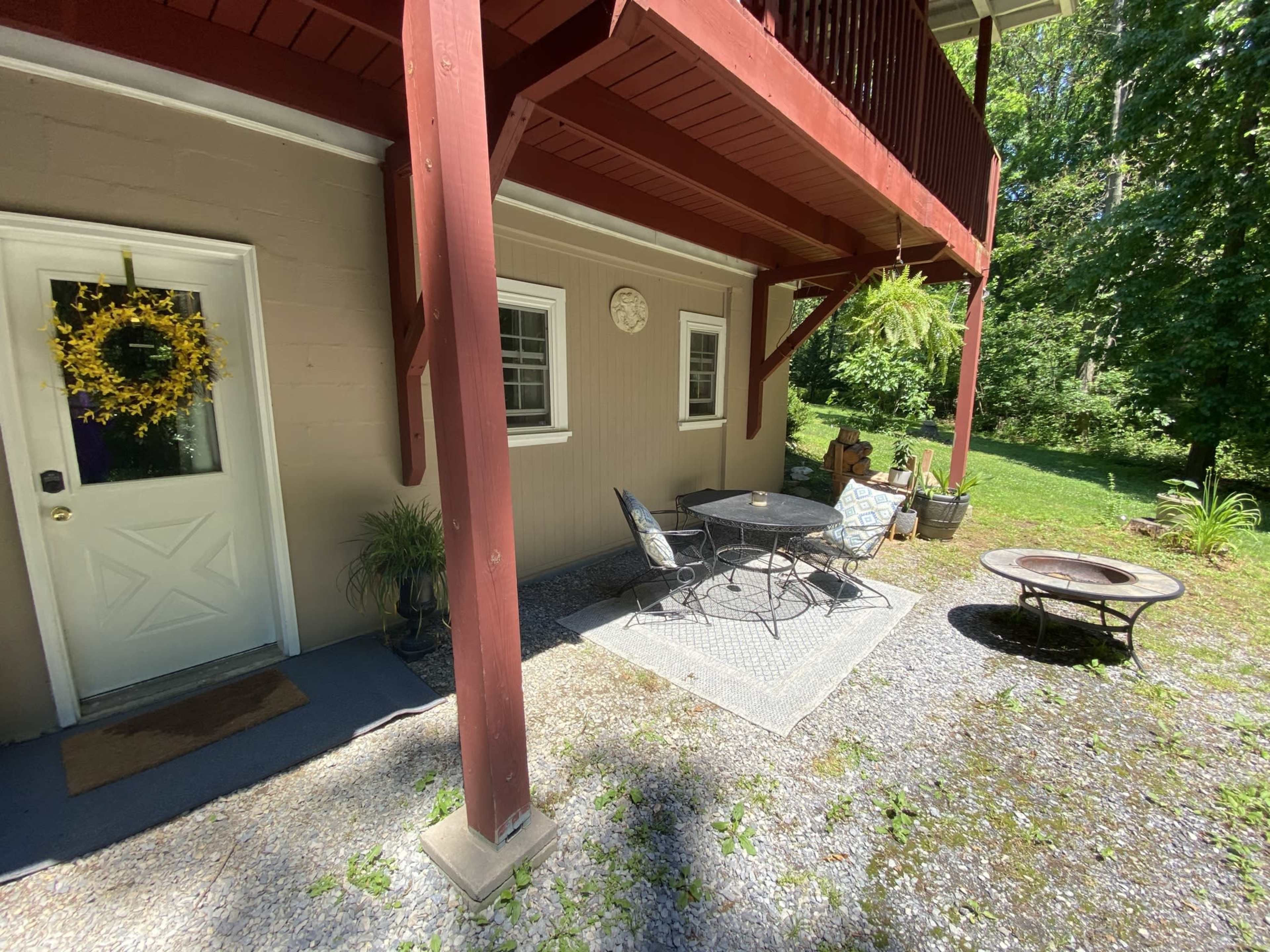 A patio area beneath a wooden balcony, featuring a small table and chairs, a fire pit, and a decorative door with a wreath.