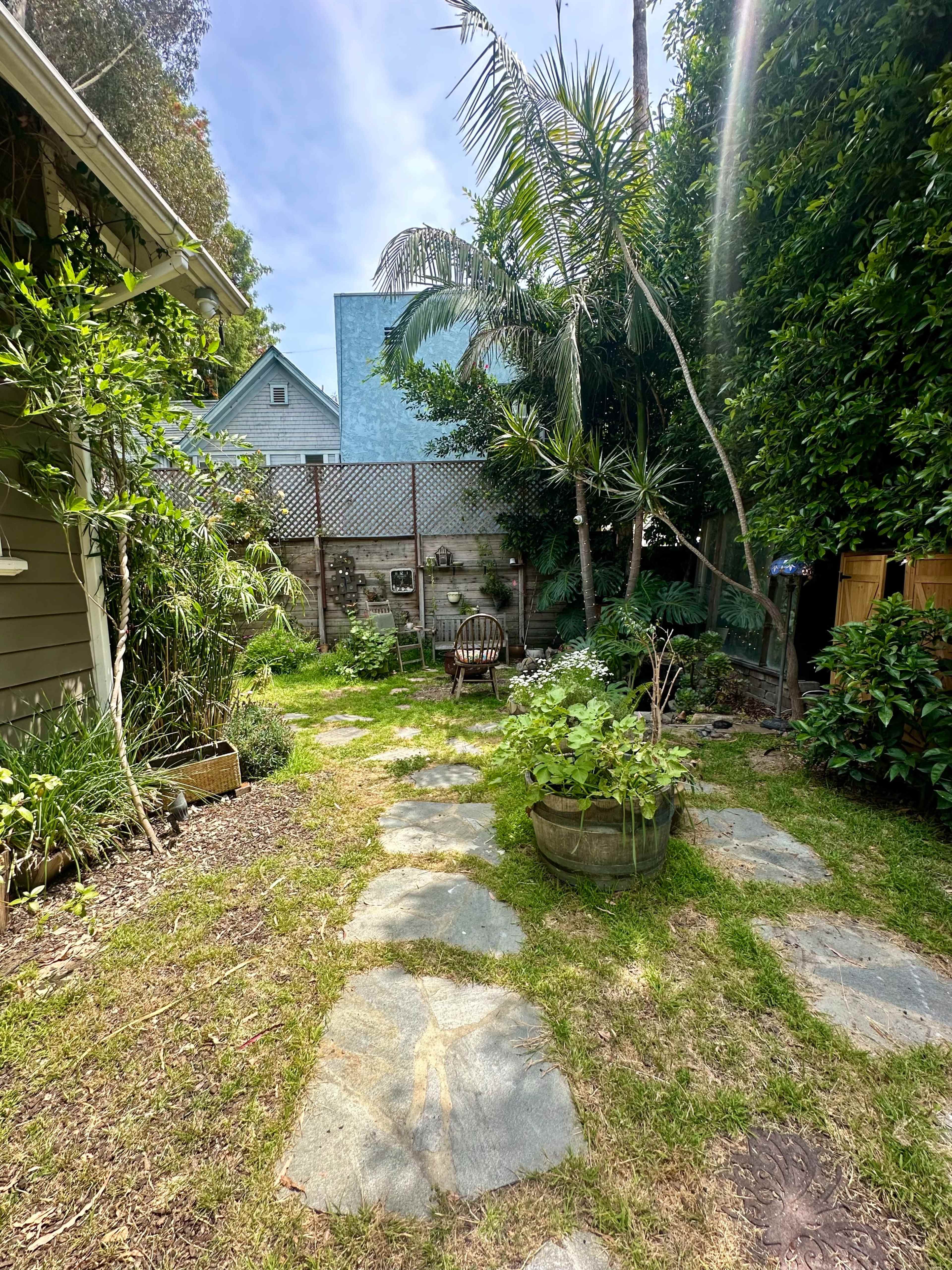 The image shows a garden with stone pathways, lush greenery, and a seating area surrounded by trees and plants.