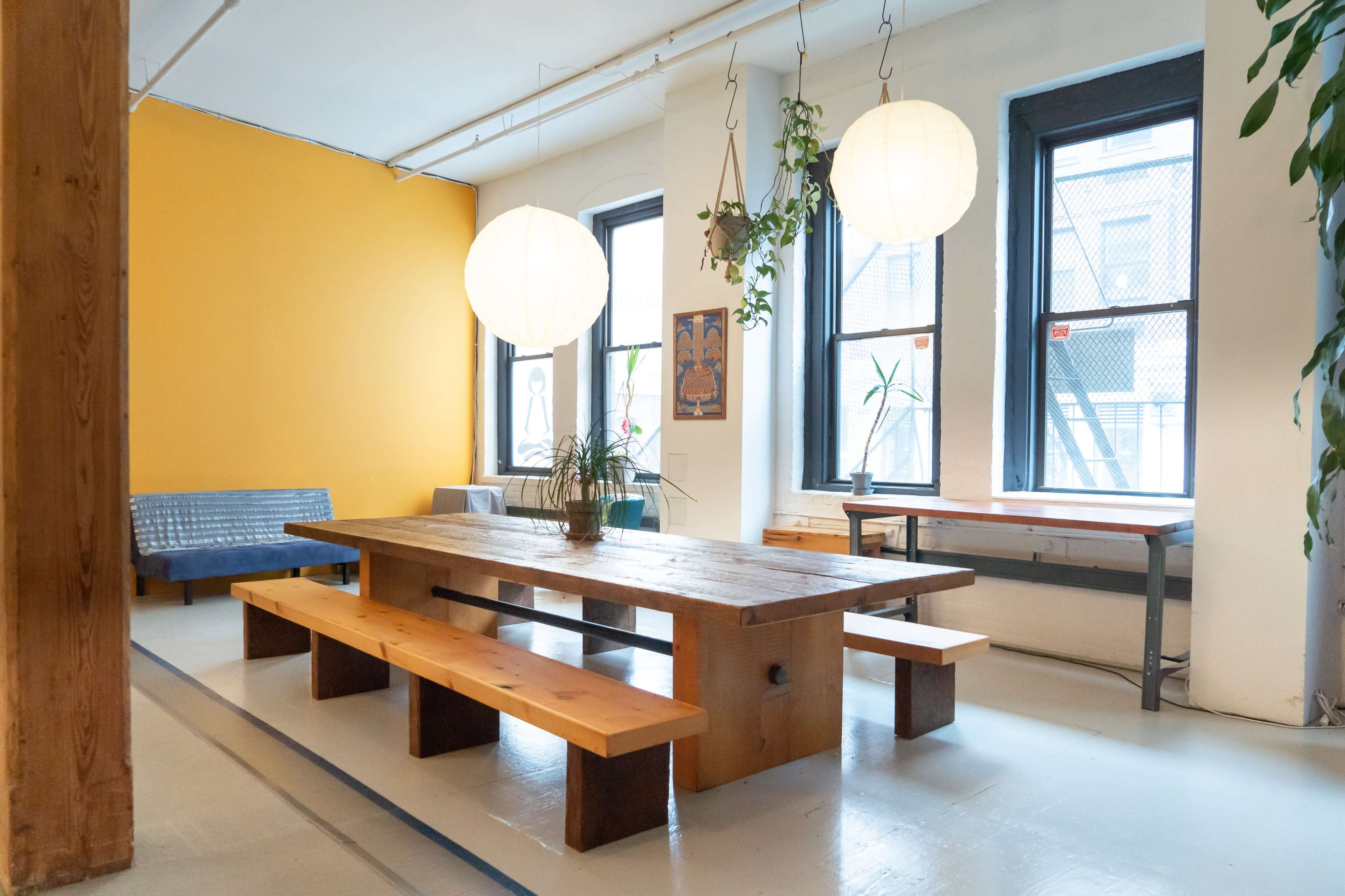 A spacious room with a large wooden table and benches surrounded by windows, featuring two hanging lanterns and a yellow wall.