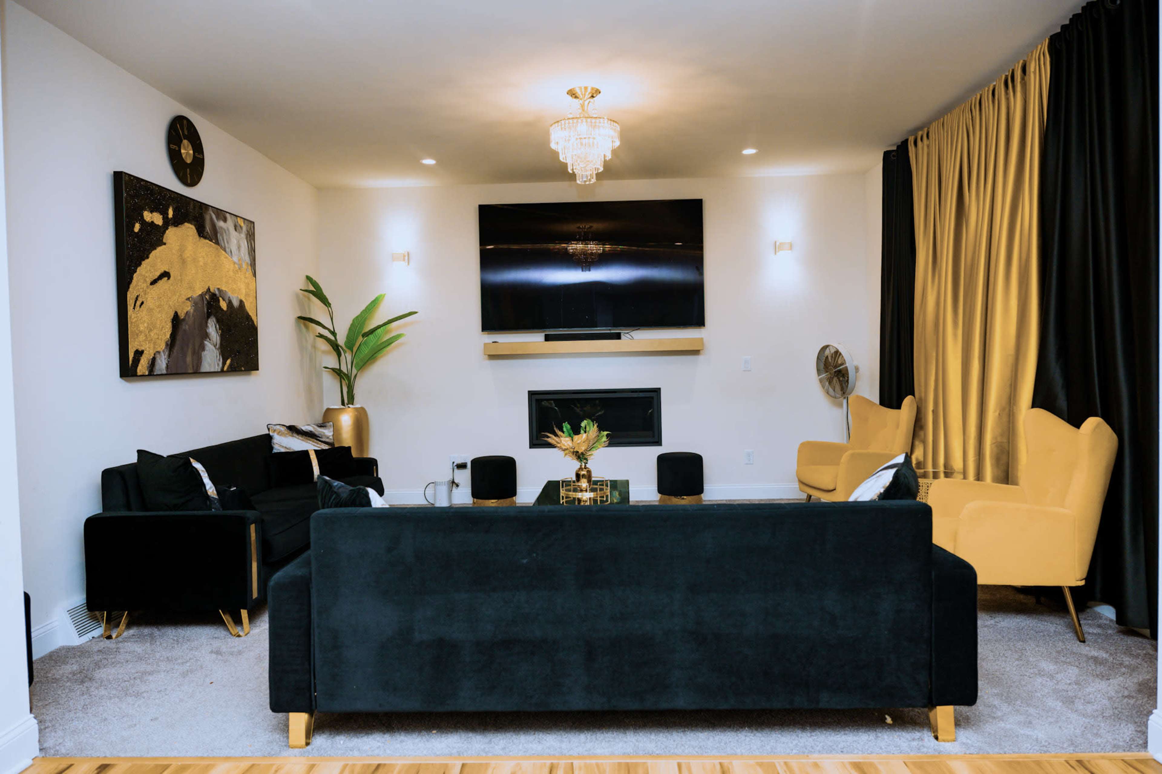 The image shows a modern living room with a large television mounted on the wall, a black sofa set, a gold-accented coffee table, and two yellow armchairs, all complemented by decorative plants and artwork.