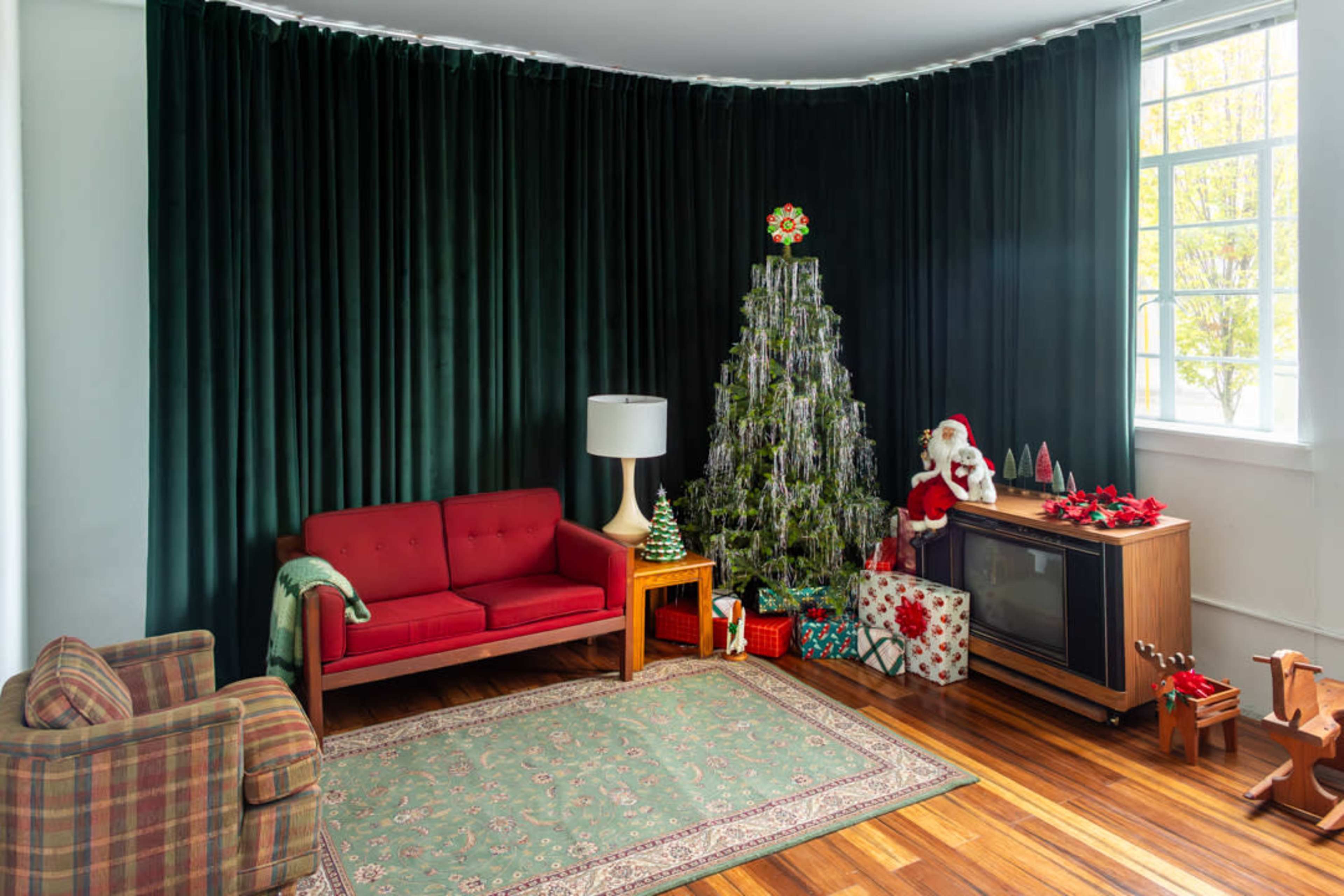 The image shows a decorated living room with a green velvet curtain, a red couch, a Christmas tree, and holiday decorations around a wooden floor.