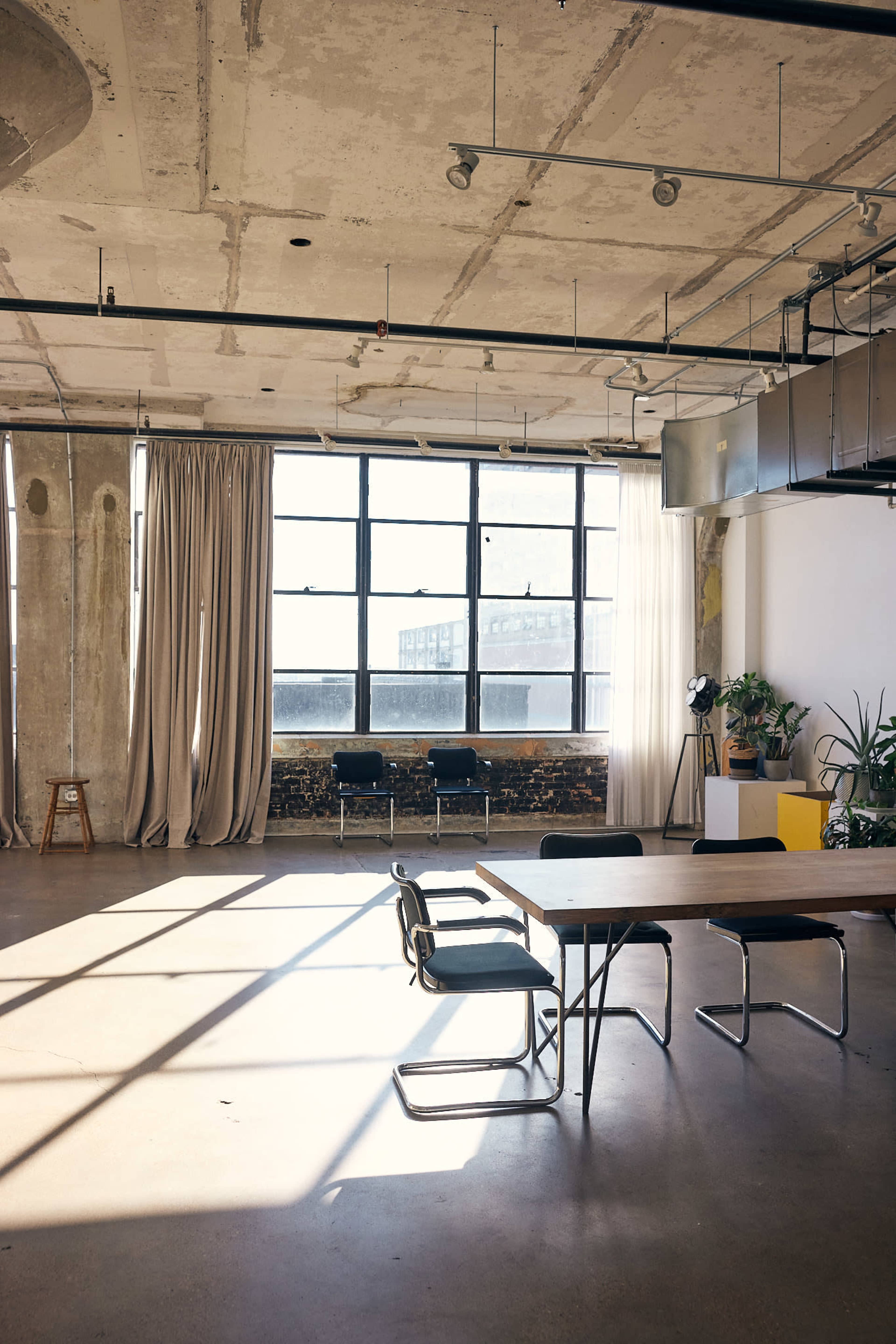The image depicts a spacious, well-lit interior of an office or studio, featuring a large window, a wooden table with chairs, and plants alongside minimalistic decor.