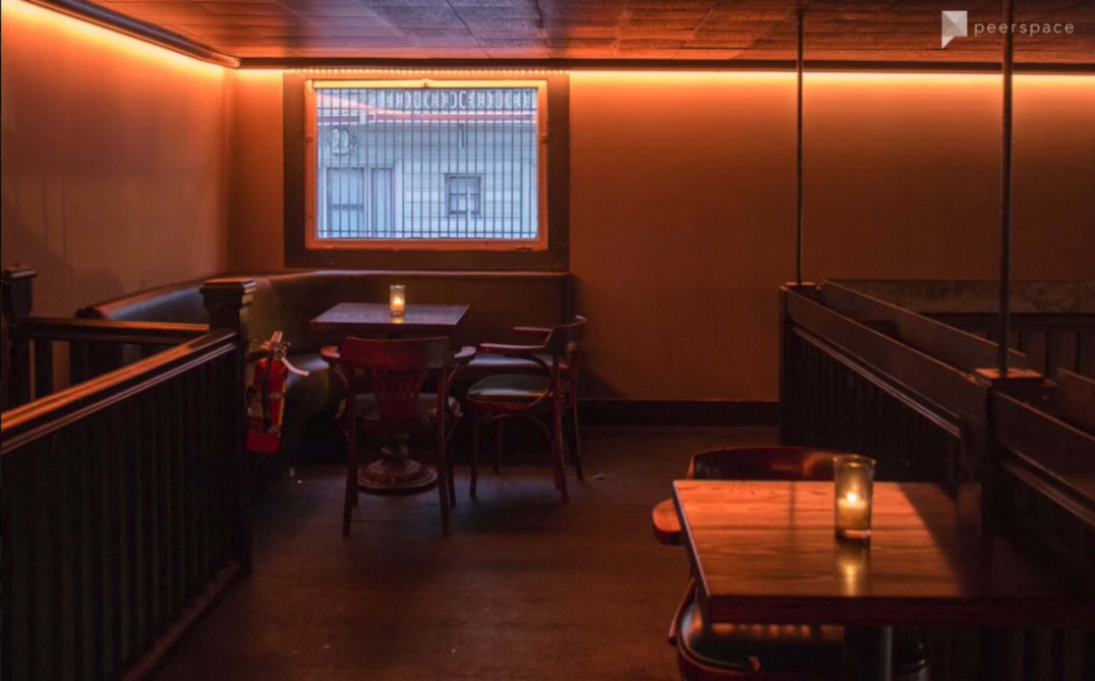 A dimly lit corner of a restaurant or bar with wooden tables, chairs, and a window featuring warm light along the walls.