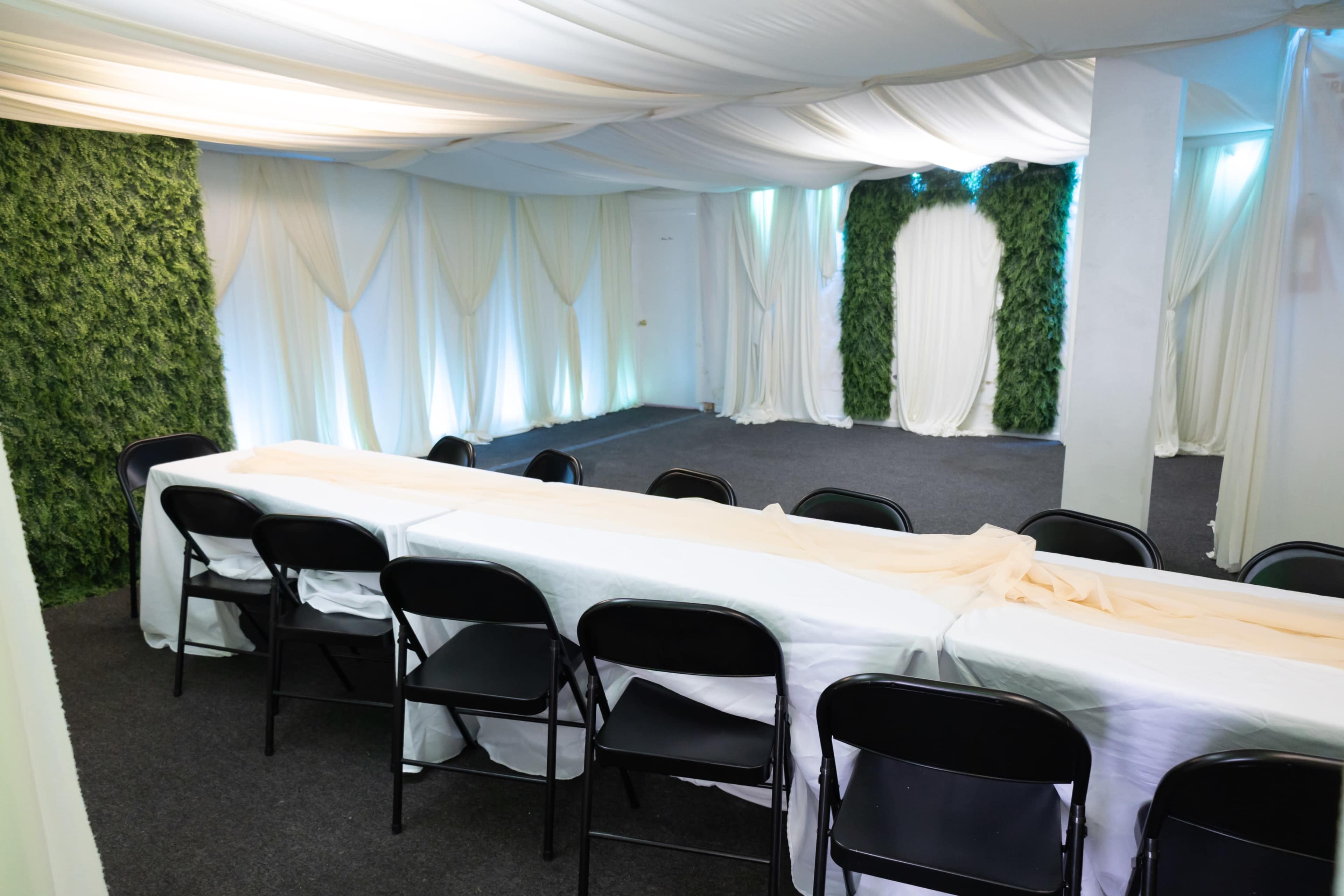 The image shows a spacious indoor venue with a long table covered in a white tablecloth and black chairs arranged around it, surrounded by walls draped in white fabric and green foliage.