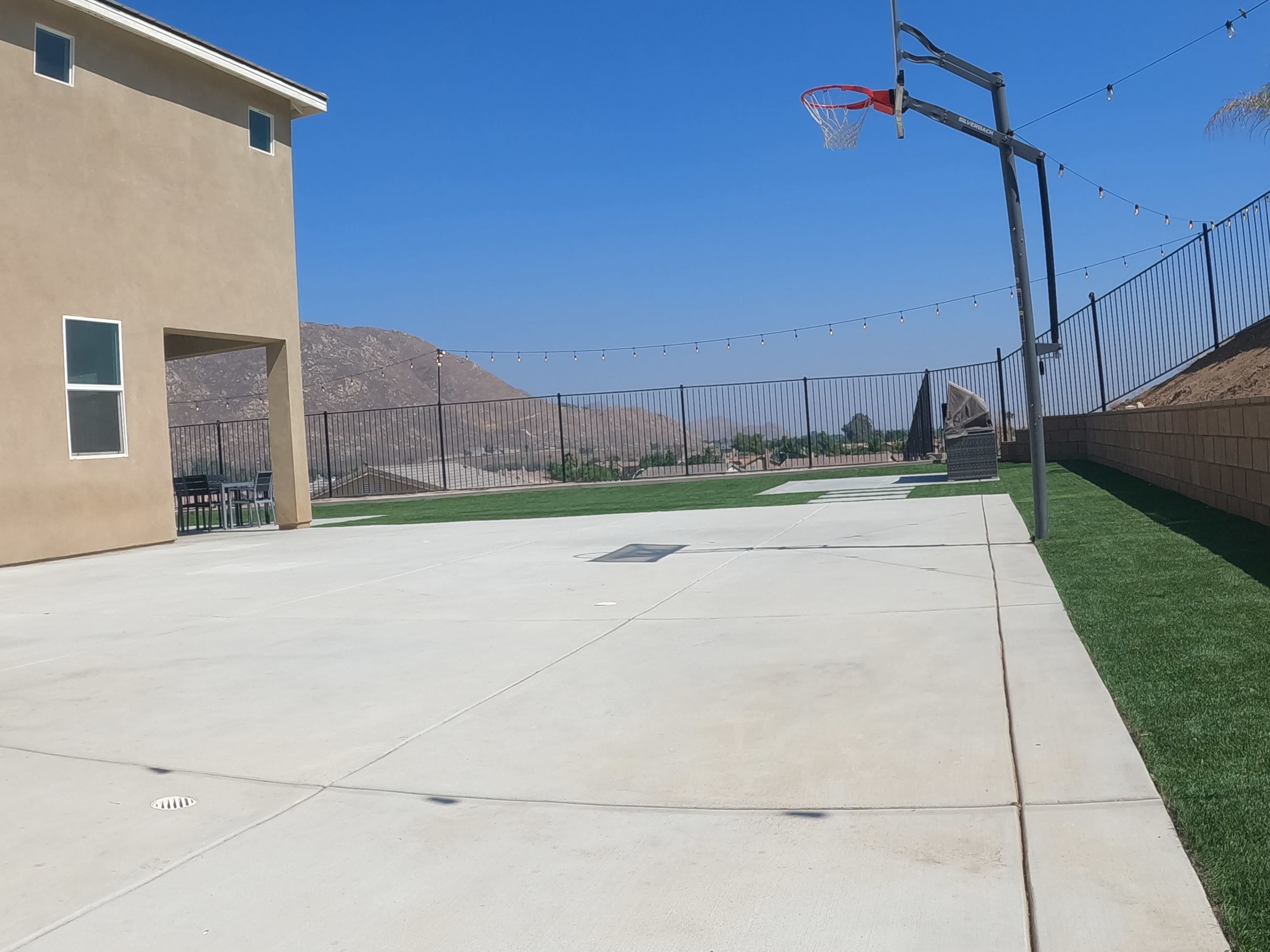 The image shows a spacious outdoor area with a basketball hoop, smooth concrete flooring, and a fenced-in landscape featuring hills in the background.
