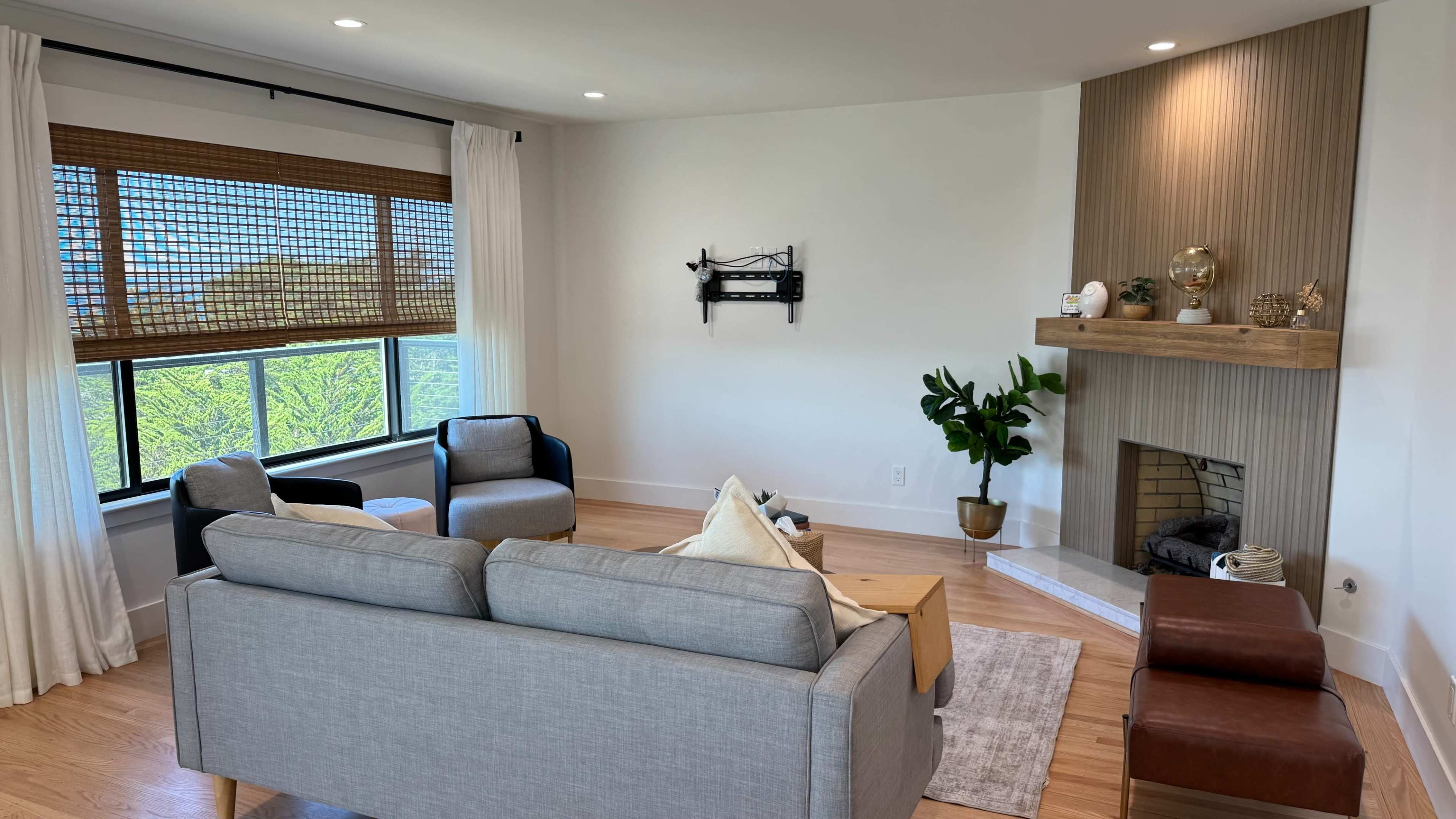 A furnished living room features a gray sofa, two black chairs, a wooden coffee table, and a plant beside a decorative fireplace with a wooden mantel.