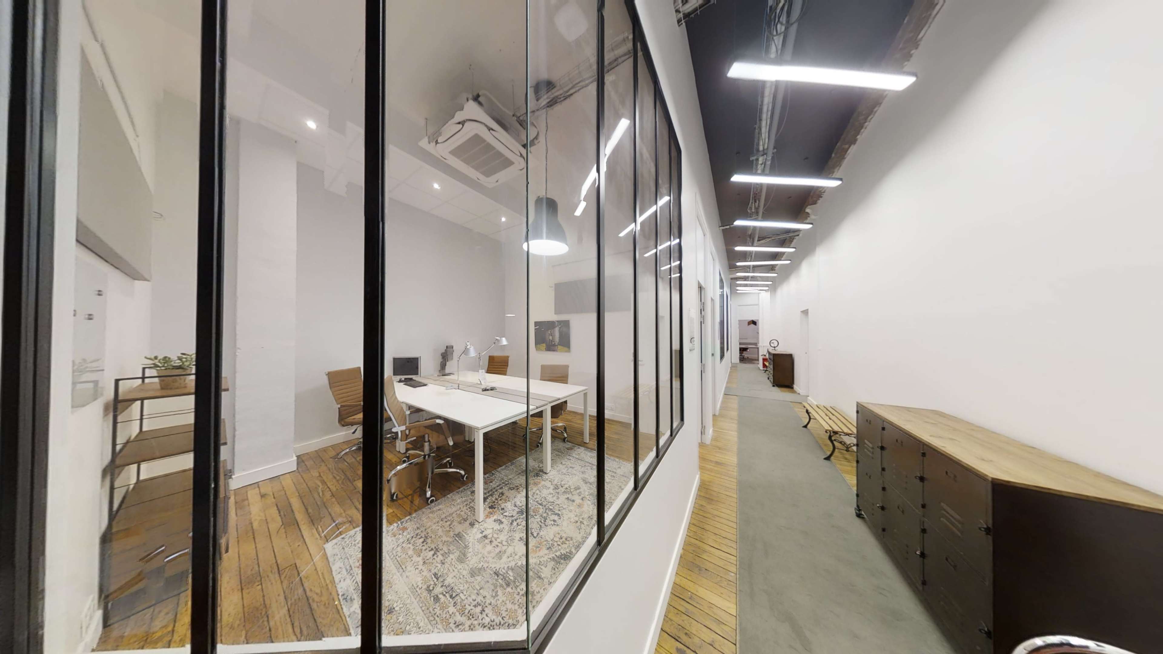 The image shows a modern office space with glass walls, featuring a meeting room with a table and chairs, and a hallway lined with wooden flooring and storage cabinets.