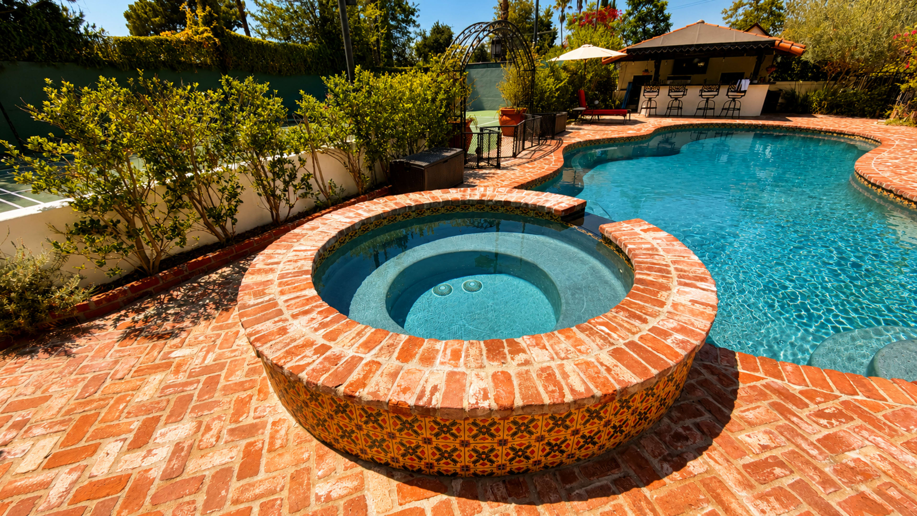 The image shows a landscaped pool area featuring a round spa integrated with a brick border and a swimming pool surrounded by greenery and a shaded seating area.