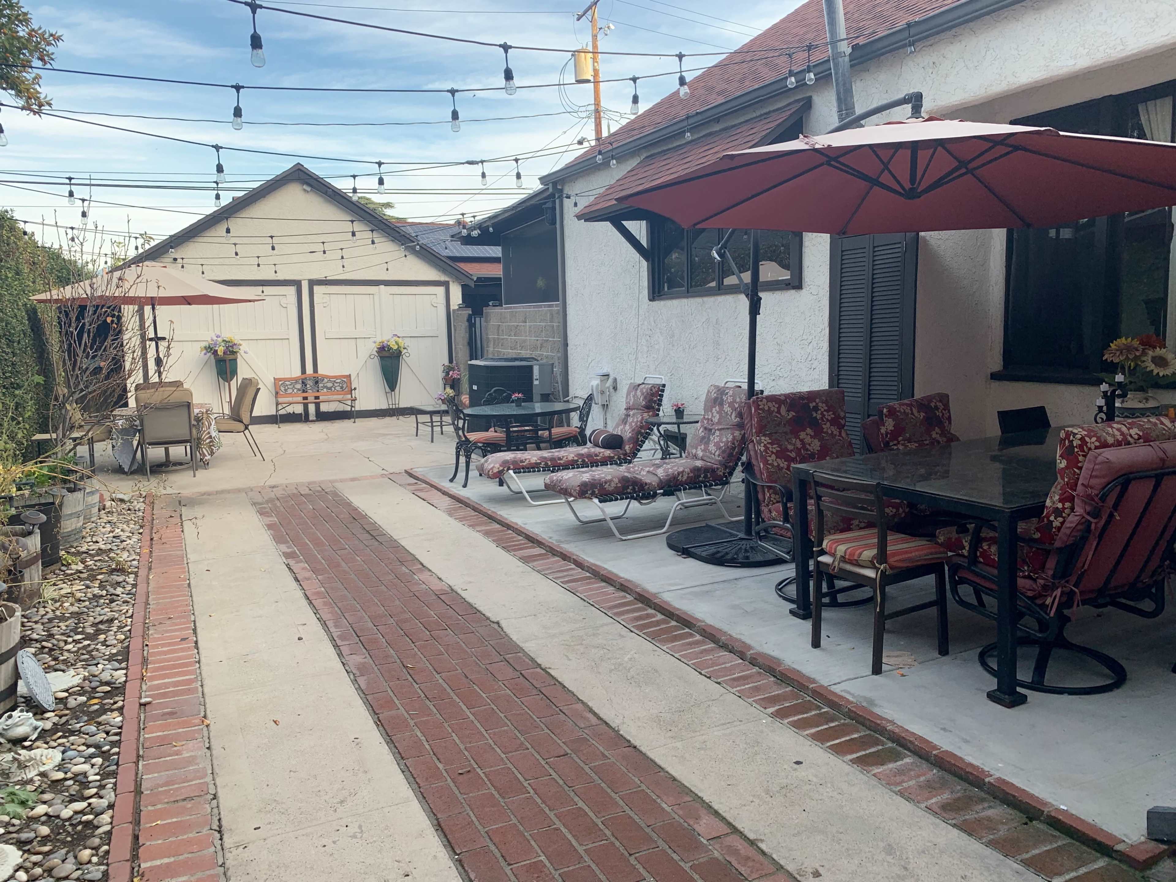The image shows a patio area with outdoor furniture, including chairs and tables, bordered by a brick pathway and string lights overhead.