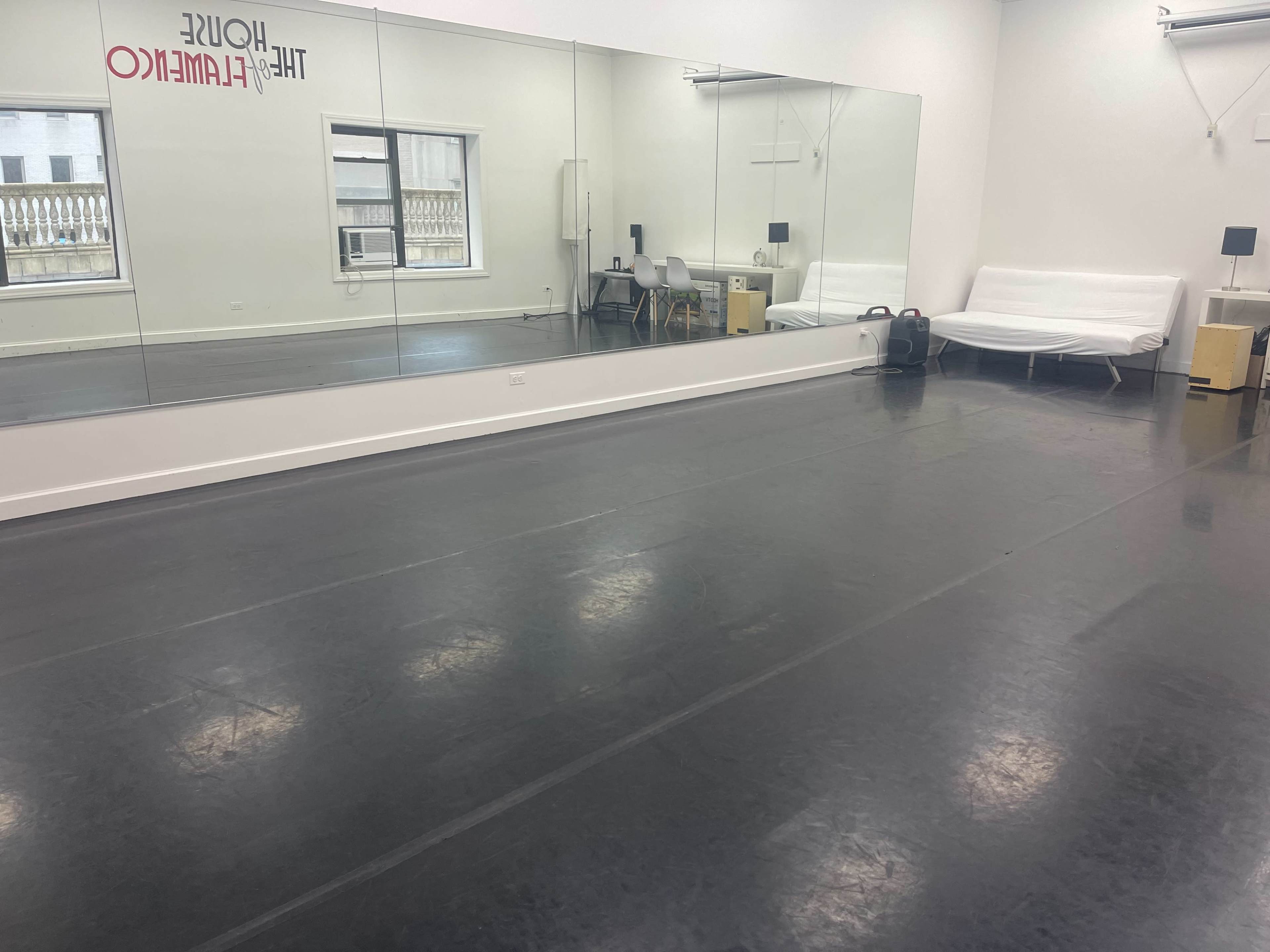The image shows a spacious dance studio with a mirrored wall, ballet flooring, and minimal furniture including a sofa and chairs.