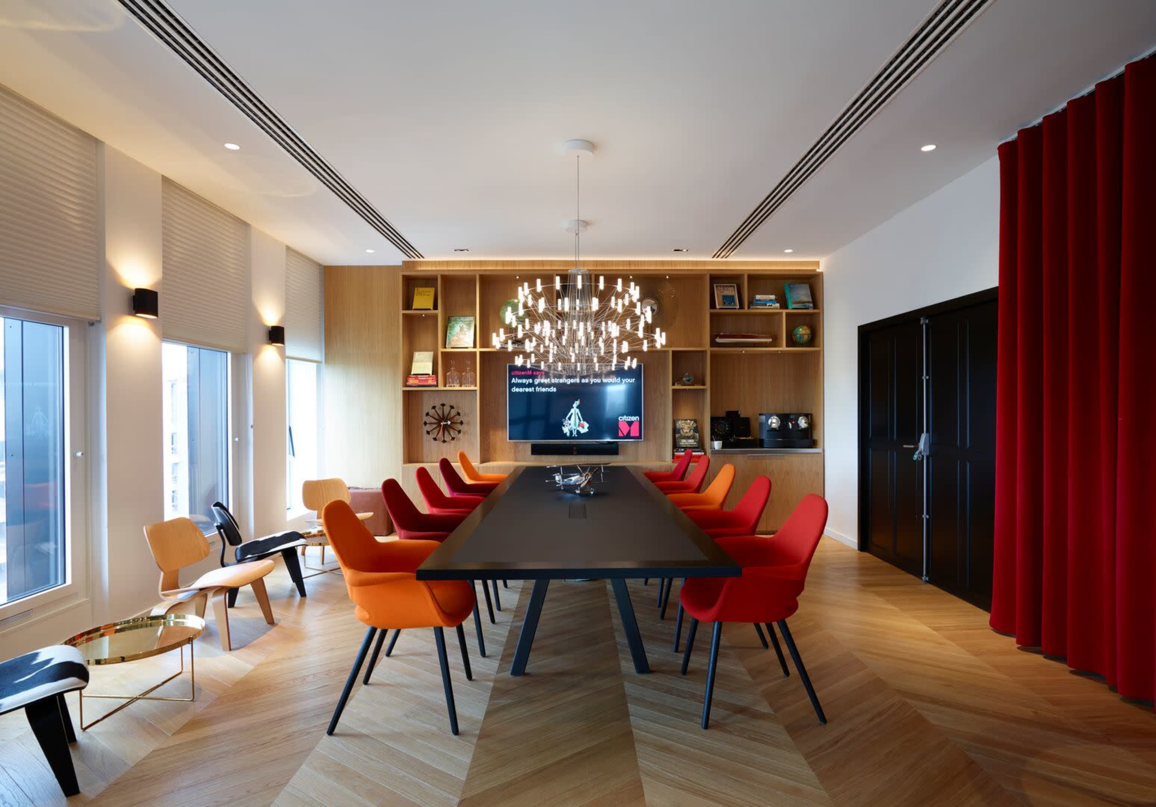A modern conference room features a large black table surrounded by orange and red chairs, a large screen on the wall, and a stylish chandelier.