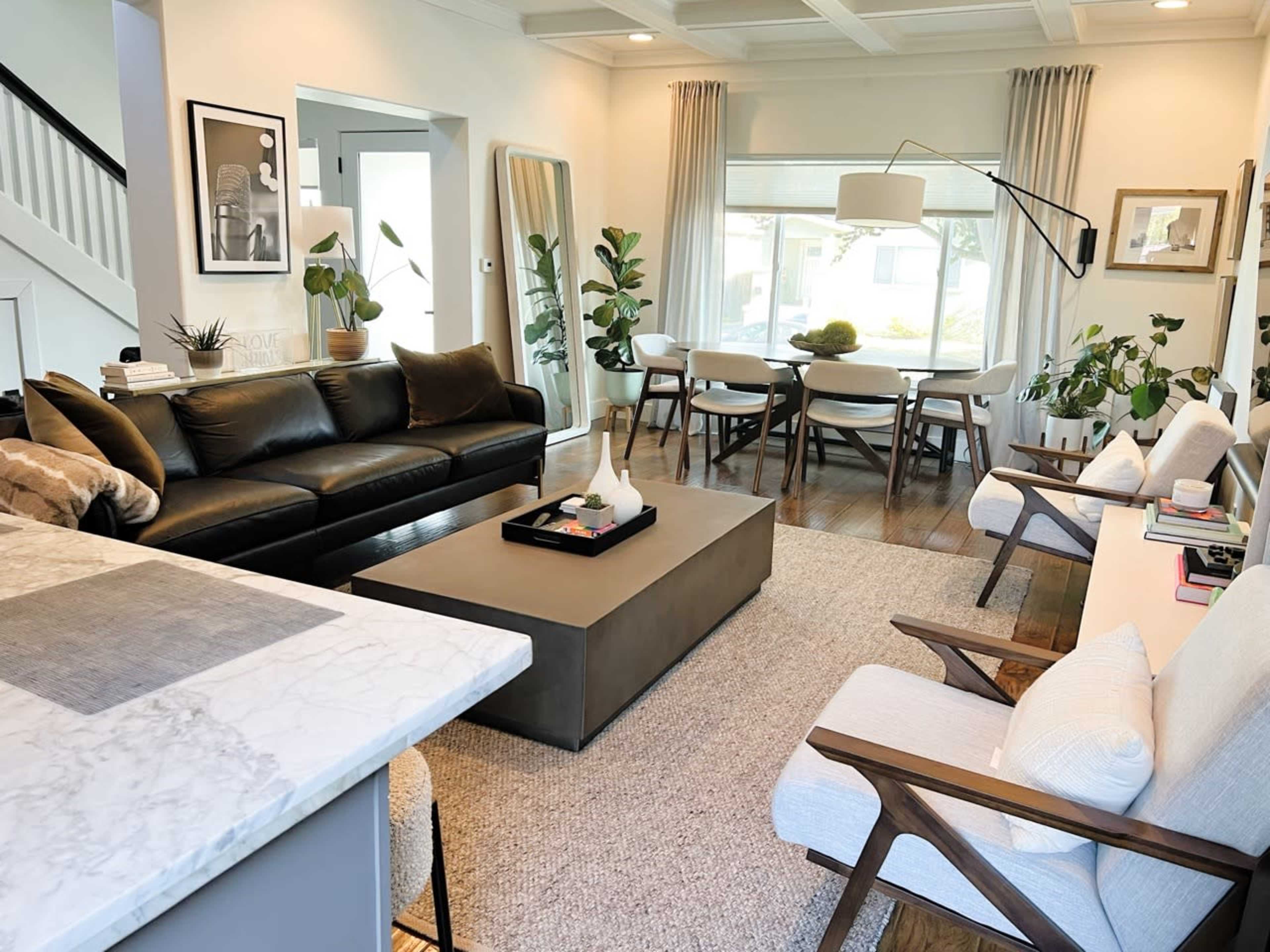 A modern living room features a black leather sofa, a coffee table, and a dining area with a large window and plants.