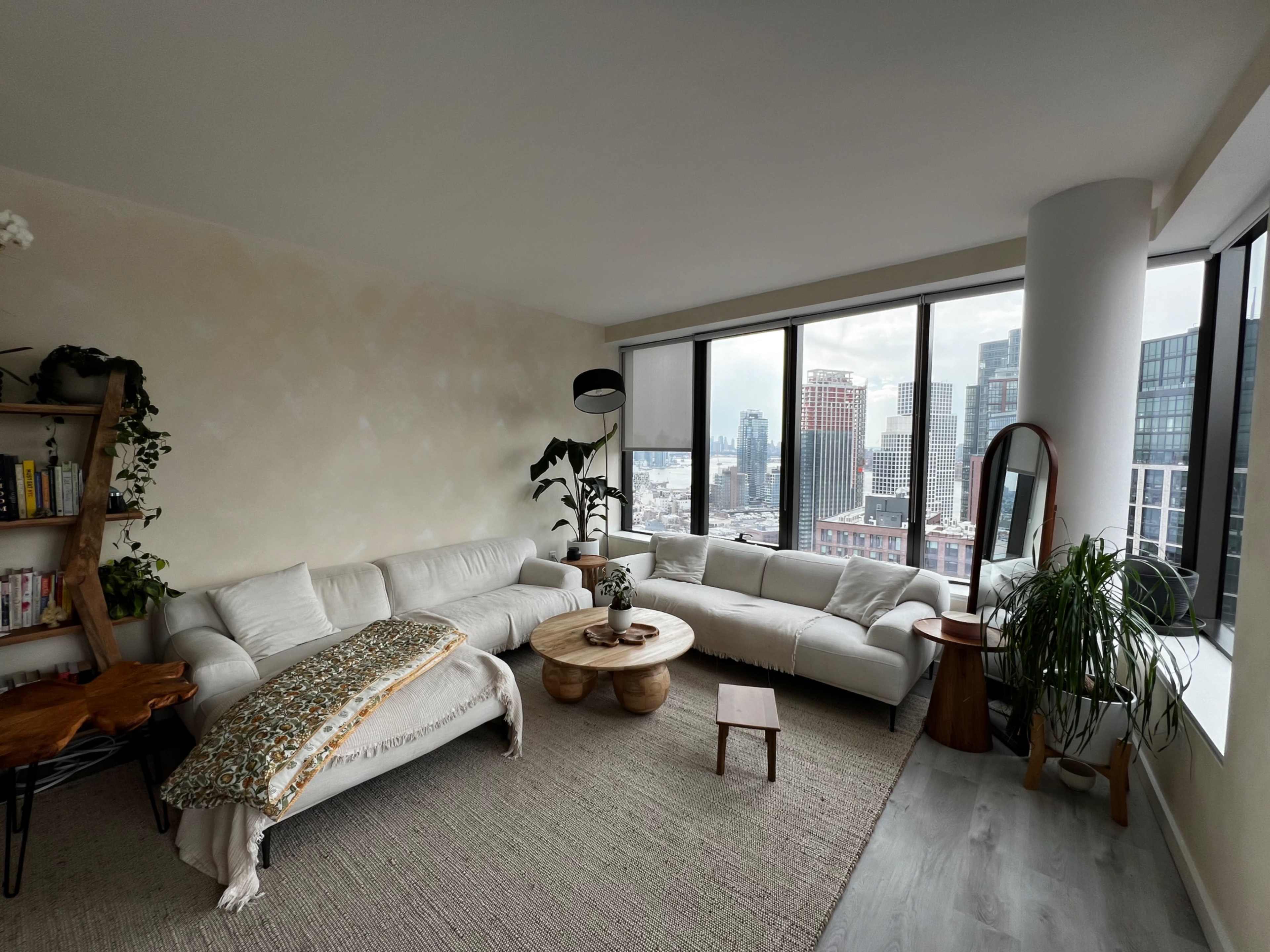 A modern living room with a white sectional sofa, a wooden coffee table, and large windows offering a view of a city skyline.