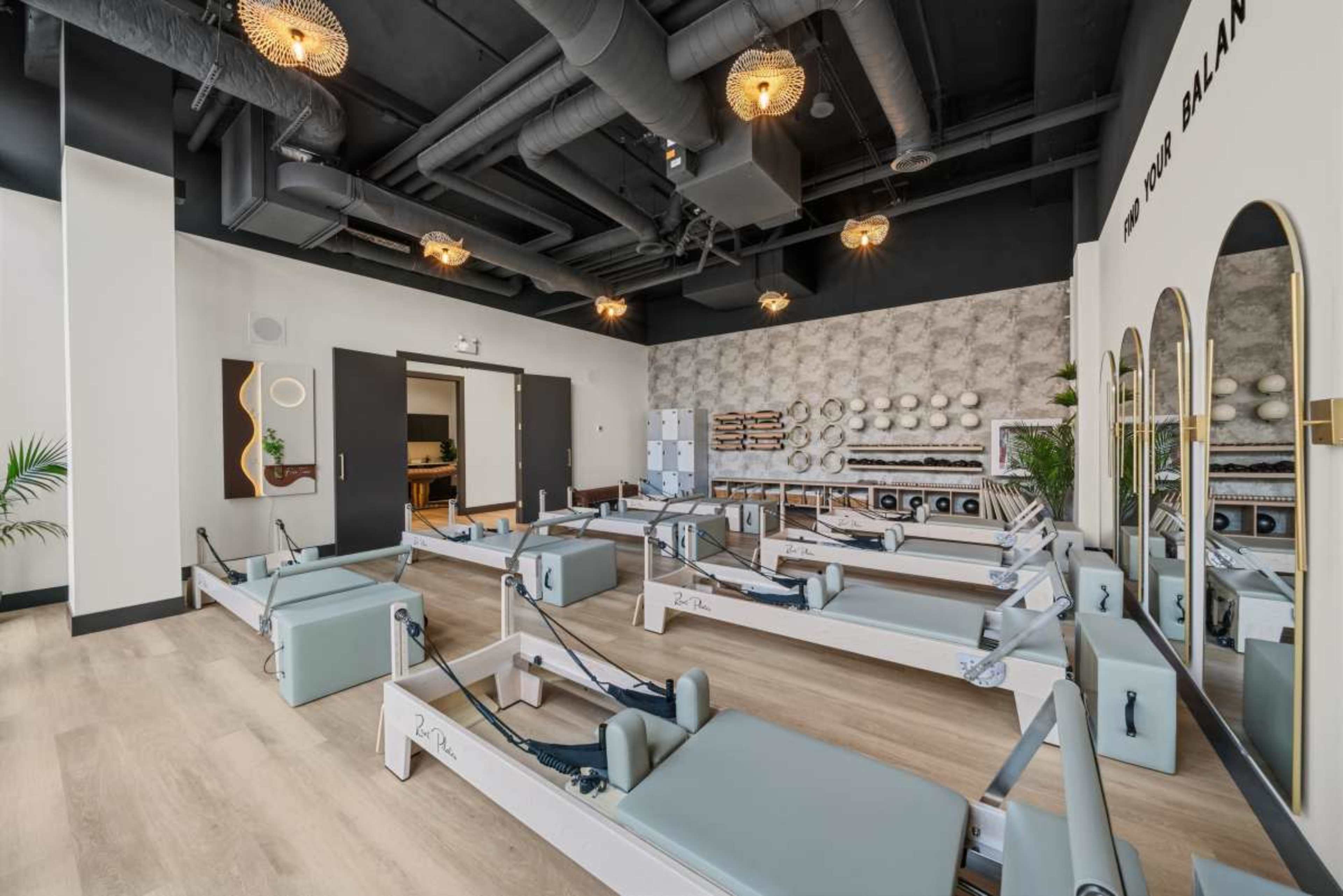 The image shows a modern pilates studio with multiple reformers arranged neatly in a well-lit space featuring decorative wall elements and stylish lighting.