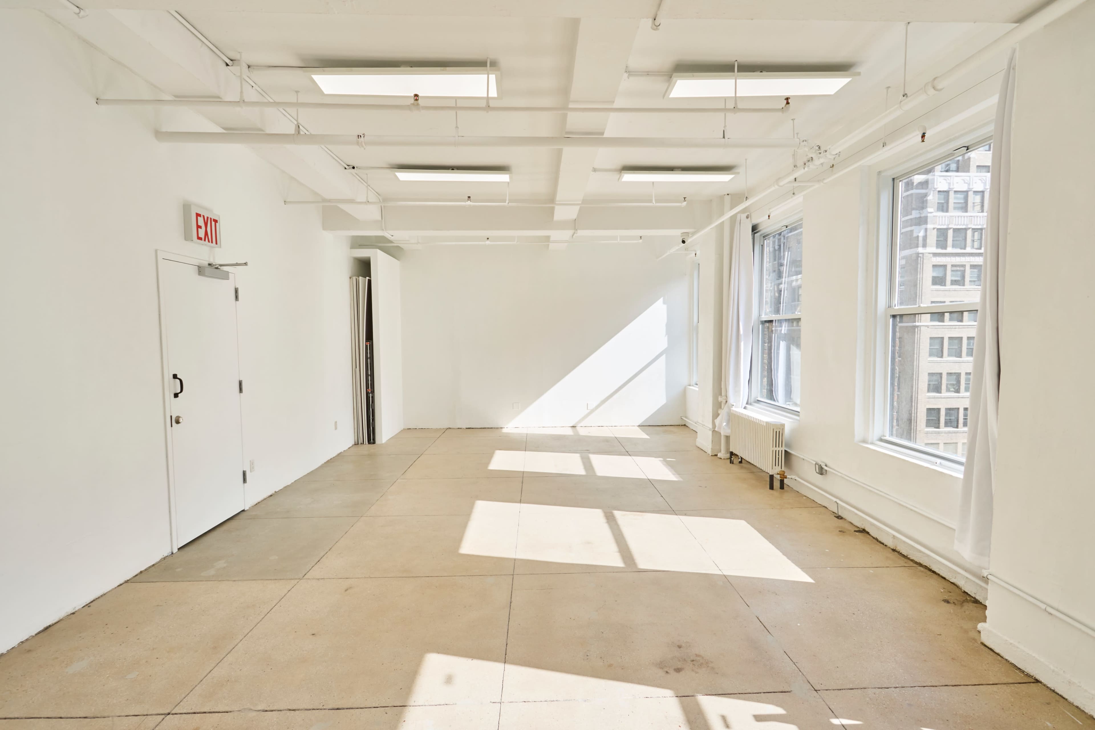The image shows a spacious, empty room with large windows allowing natural light to cast shadows on the floor.