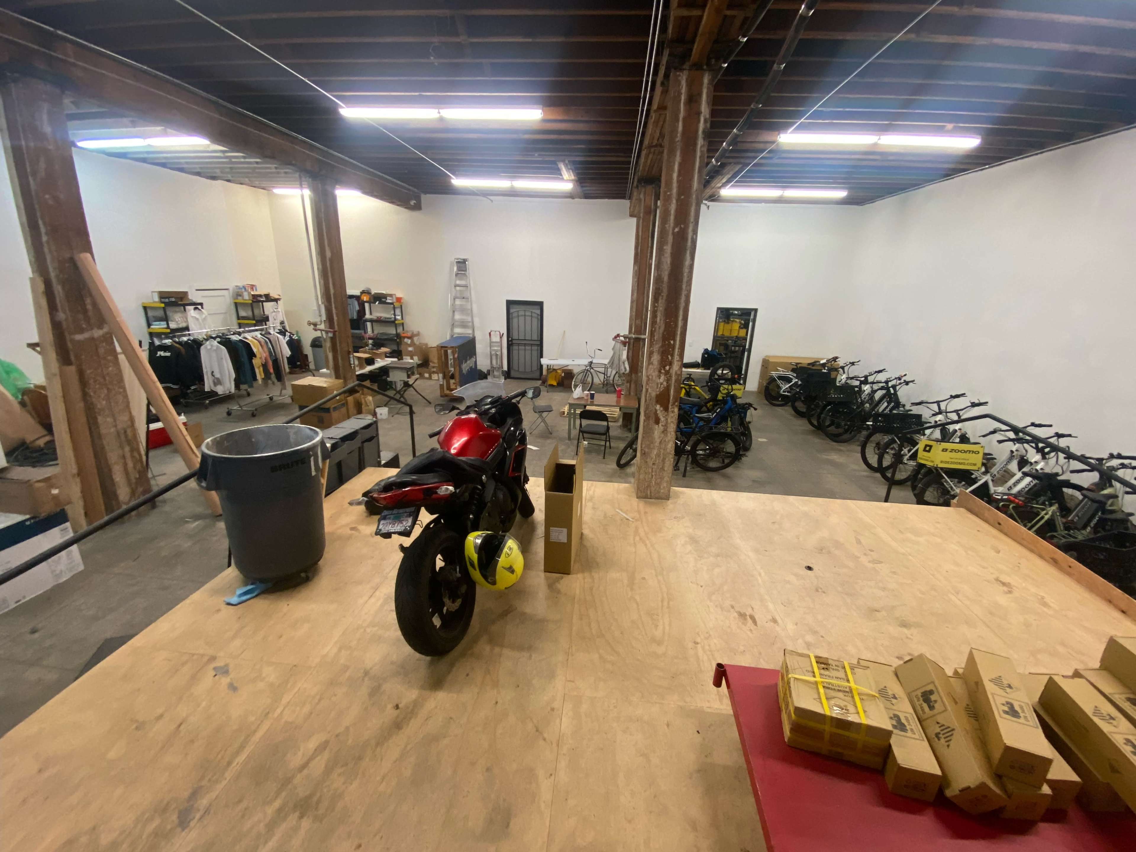 A spacious warehouse with a motorcycle on a raised wooden platform, surrounded by rows of bicycles, clothing racks, and boxes.