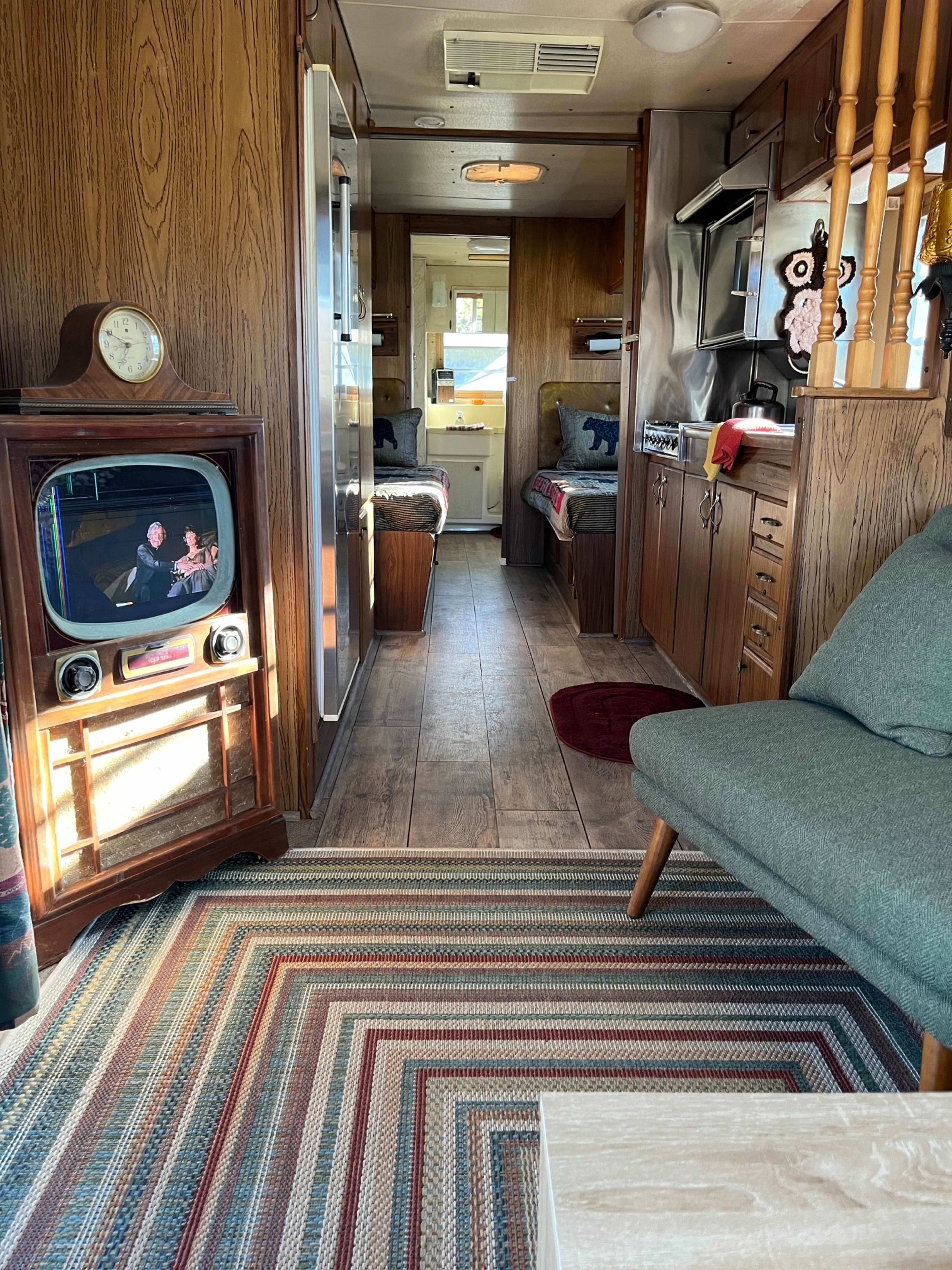The interior of a vintage camper features a living area with a retro television set, a seating area, and a kitchen space lined with wooden cabinetry.