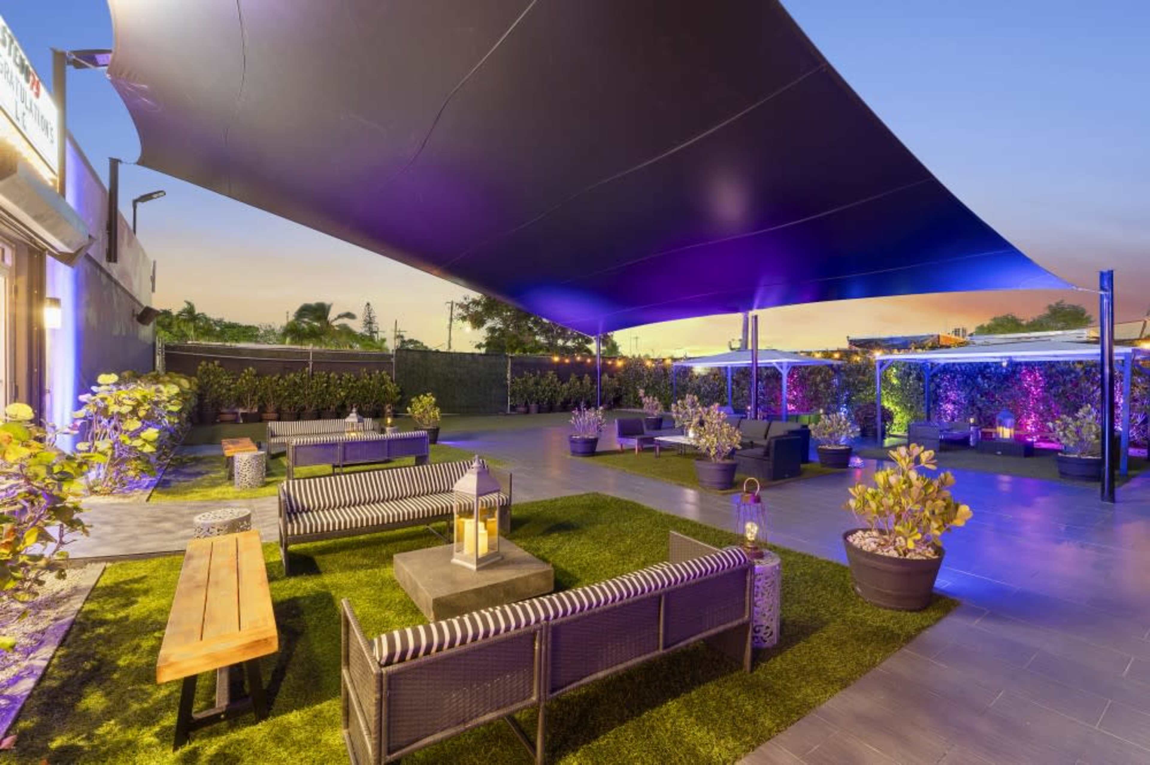 The scene features an outdoor lounge area with modern seating, surrounded by potted plants and illuminated by colorful lights under a large canopy at sunset.