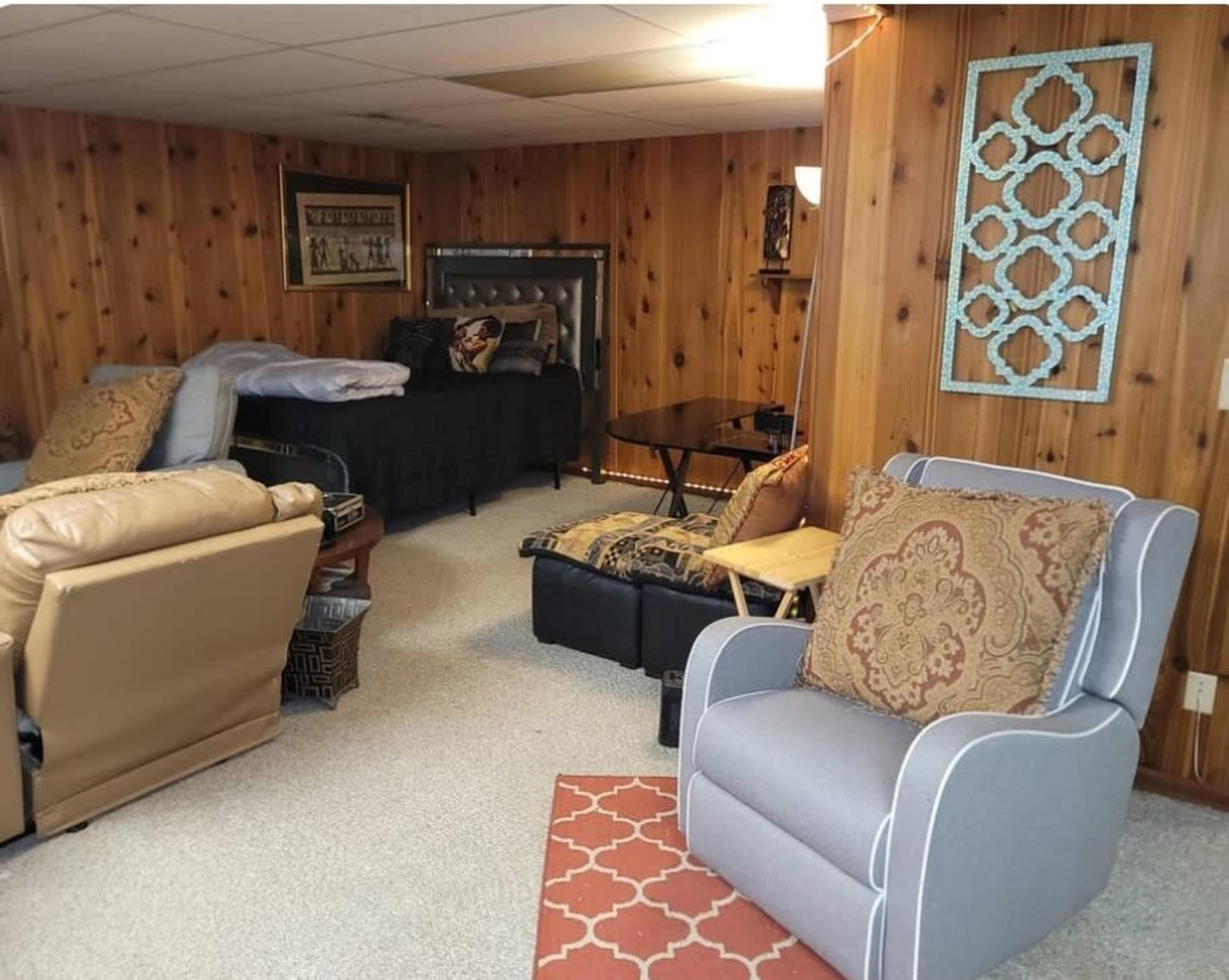 The image shows a cozy basement living area with wooden paneling, featuring a bed, a black seating area, a small table, and various upholstered chairs.