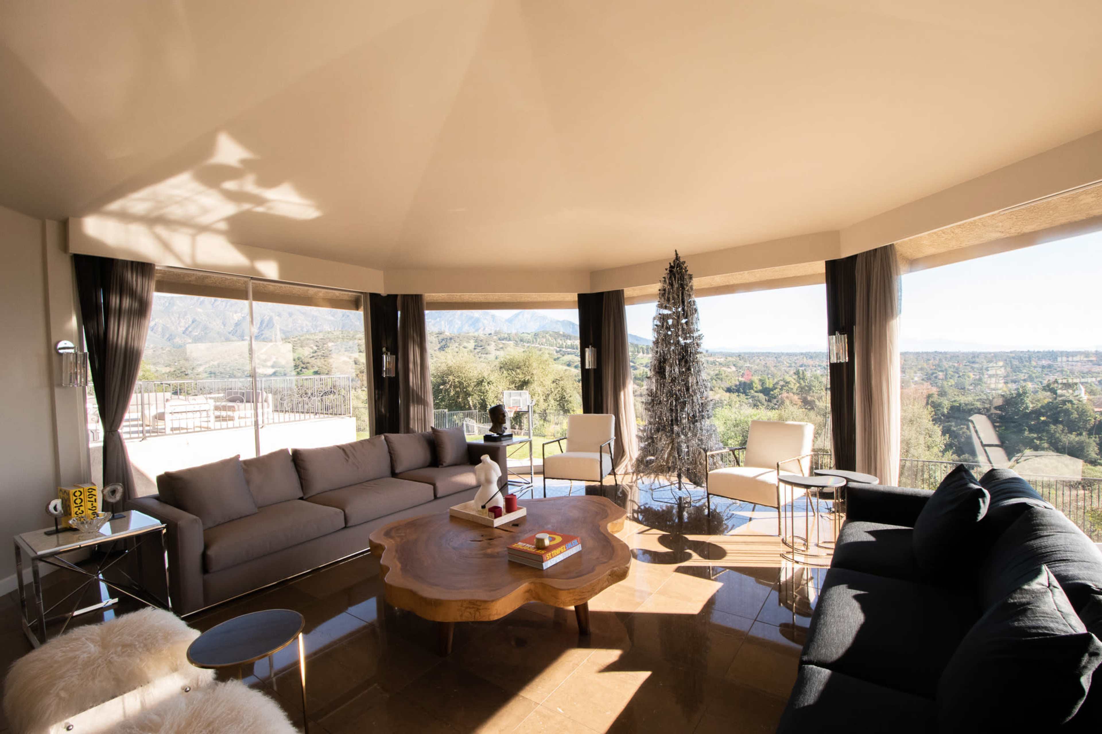 The image shows a modern living room with large windows overlooking a mountainous landscape and a Christmas tree in the corner.
