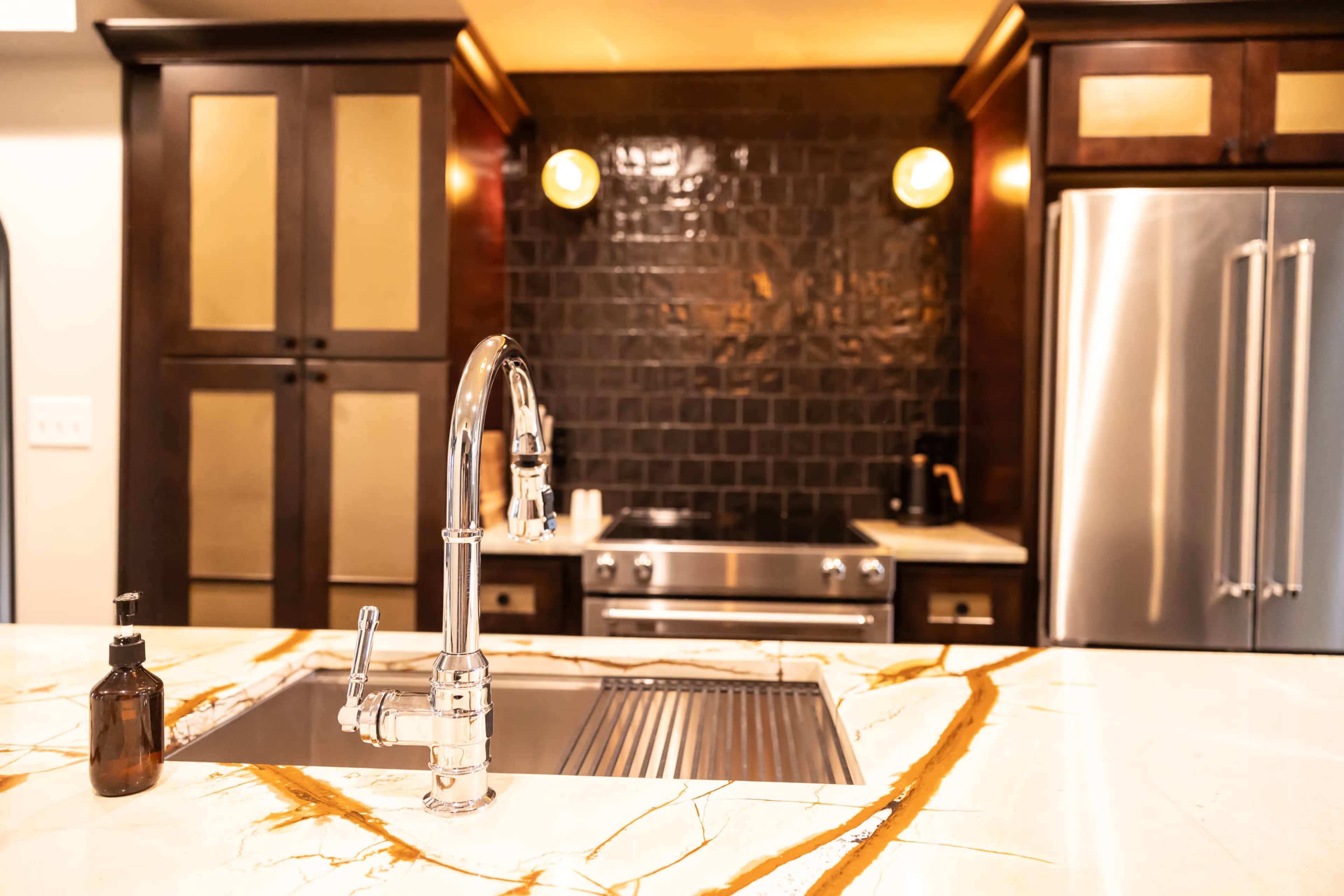 The image shows a modern kitchen with a stainless steel sink, a sleek faucet, a dark tiled backsplash, and a stove, all complemented by a marble countertop with golden veining.