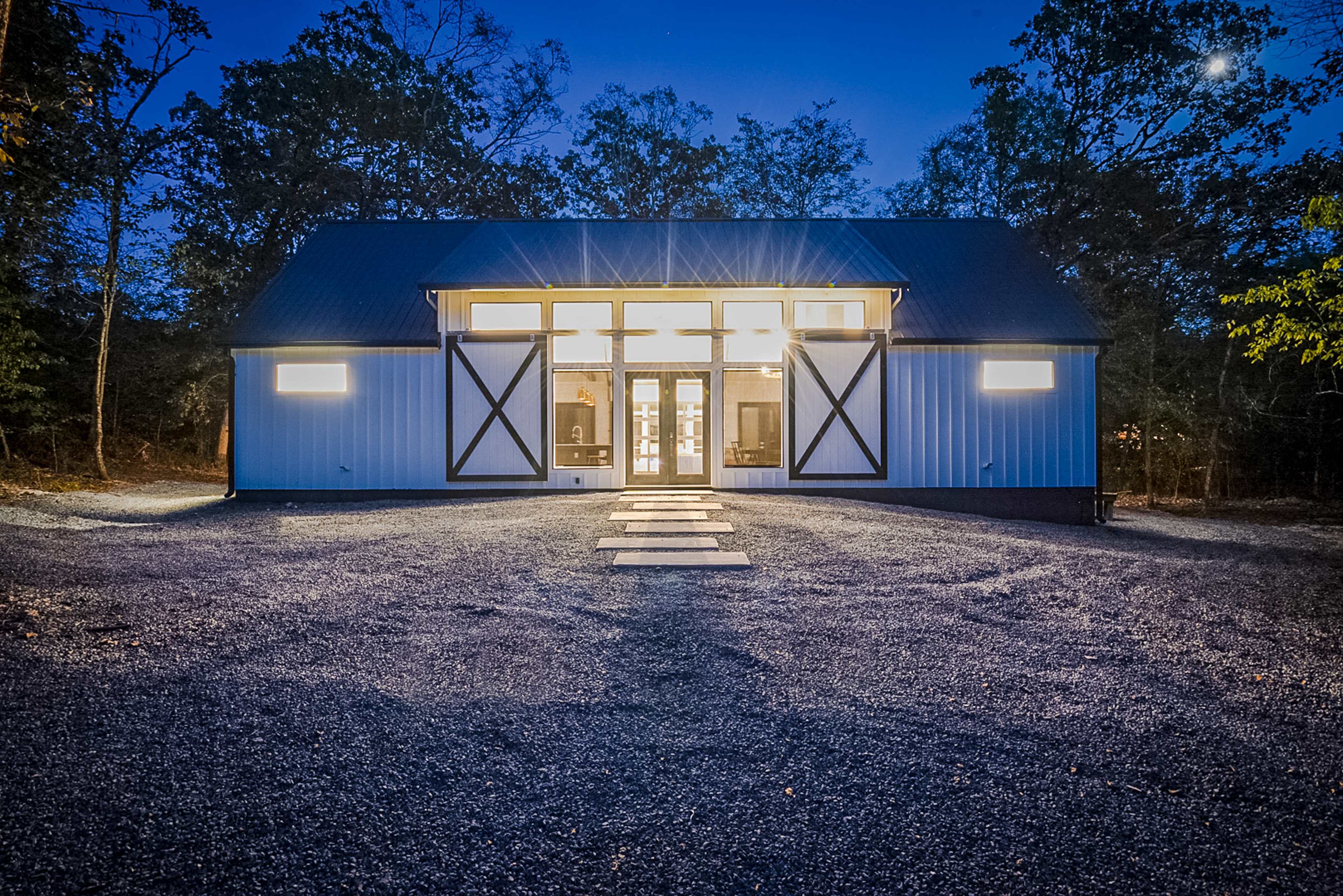 A modern barn-style building is illuminated by large windows at dusk, with a gravel pathway leading to its entrance.