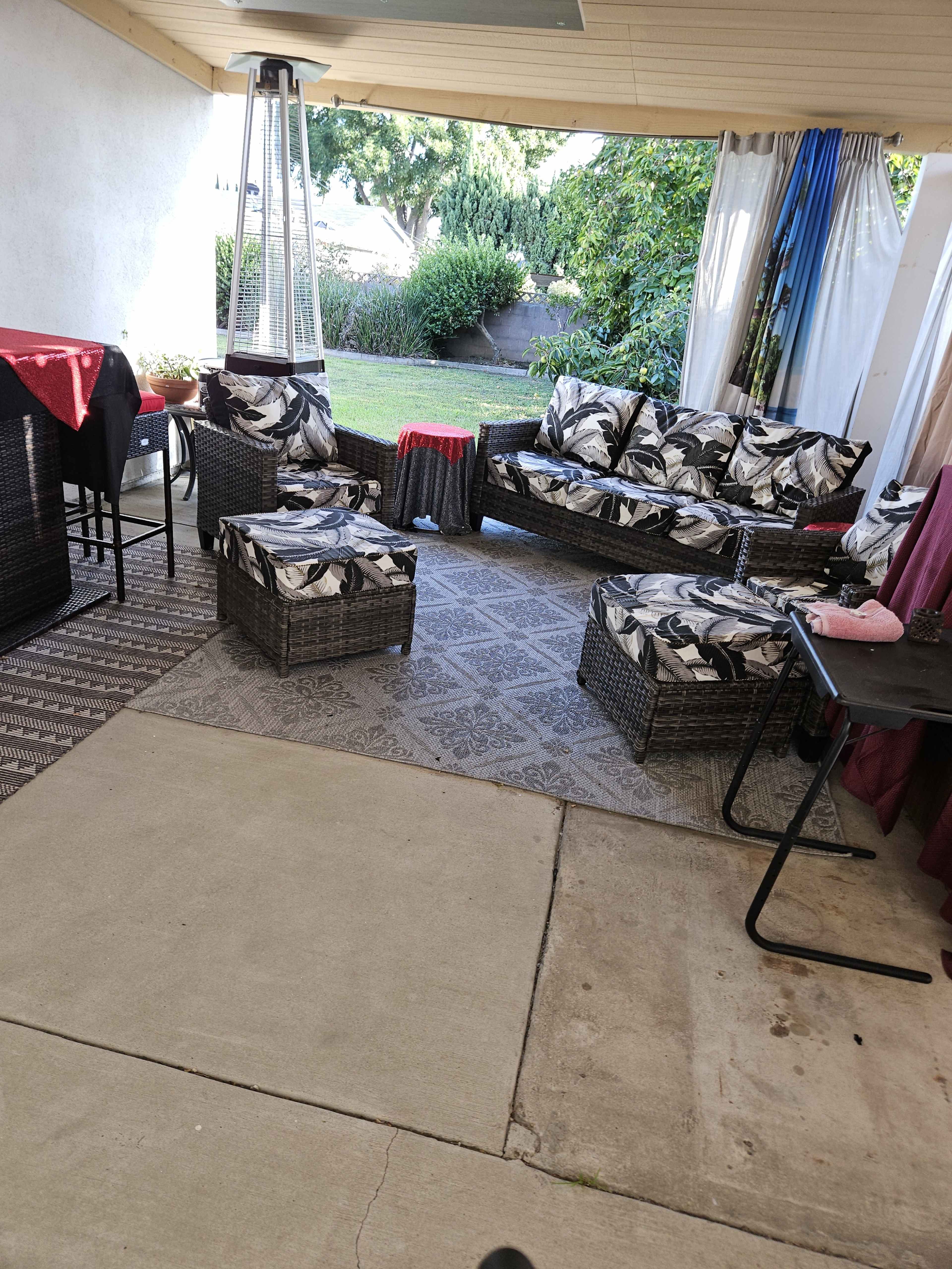 A covered outdoor seating area with wicker furniture, including a couch and several ottomans, arranged on a patio with a concrete floor.