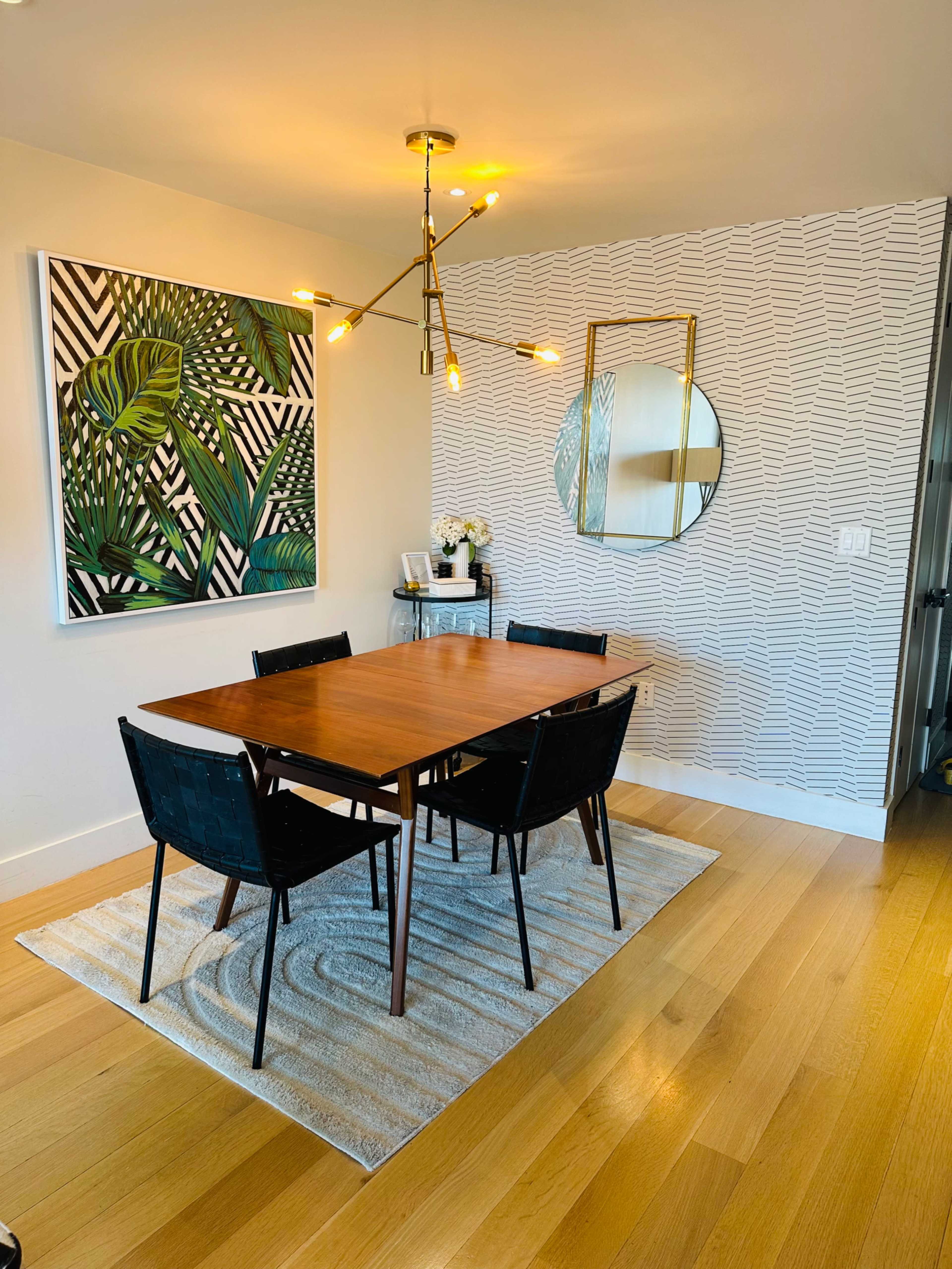 A dining room features a wooden table set with black chairs, a large artwork on the wall, and a decorative mirror reflecting the space.