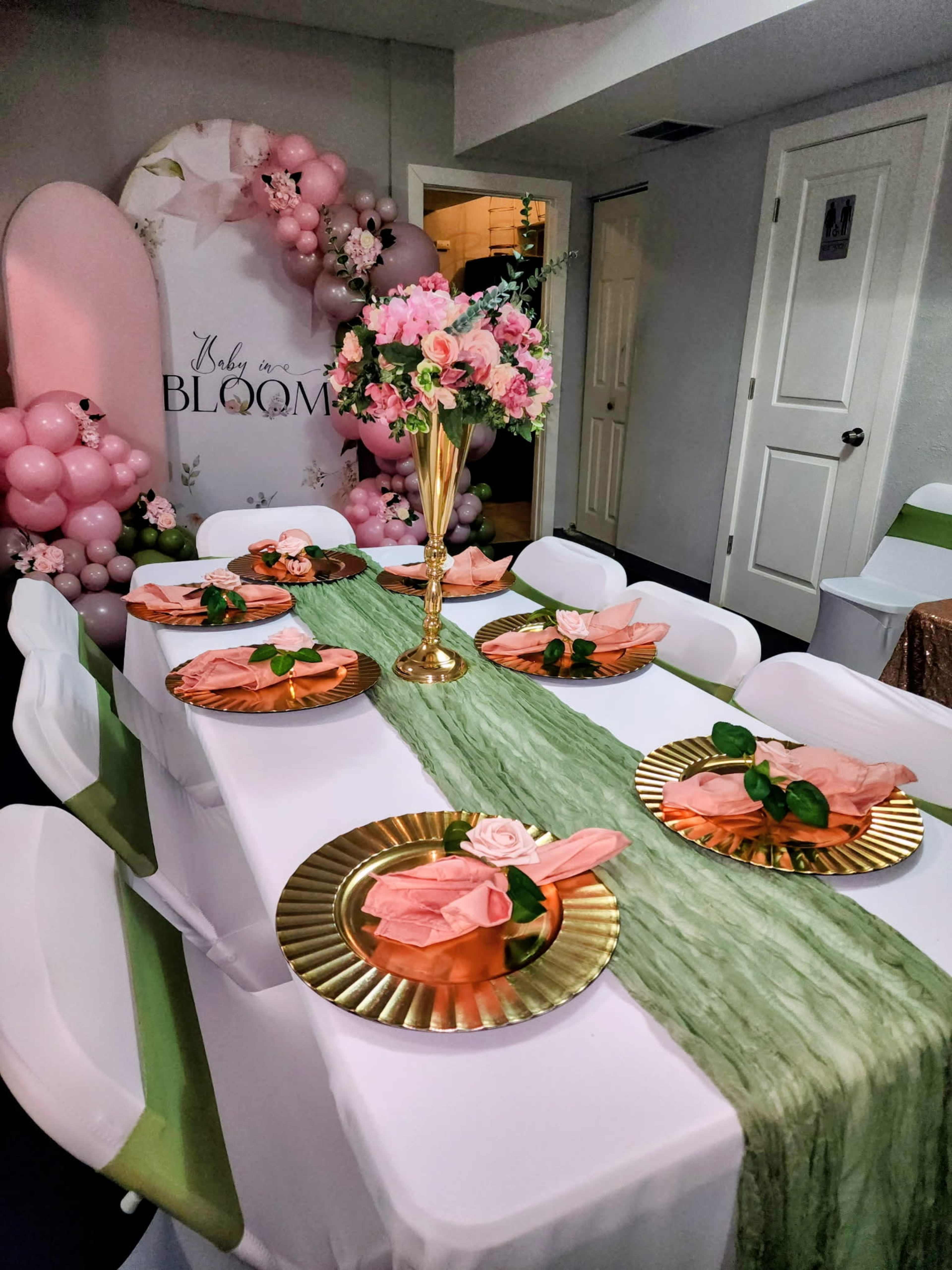 The image shows a decorated indoor space with a long dining table set with gold plates, pink napkins, and green table runners, surrounded by balloons and floral arrangements.