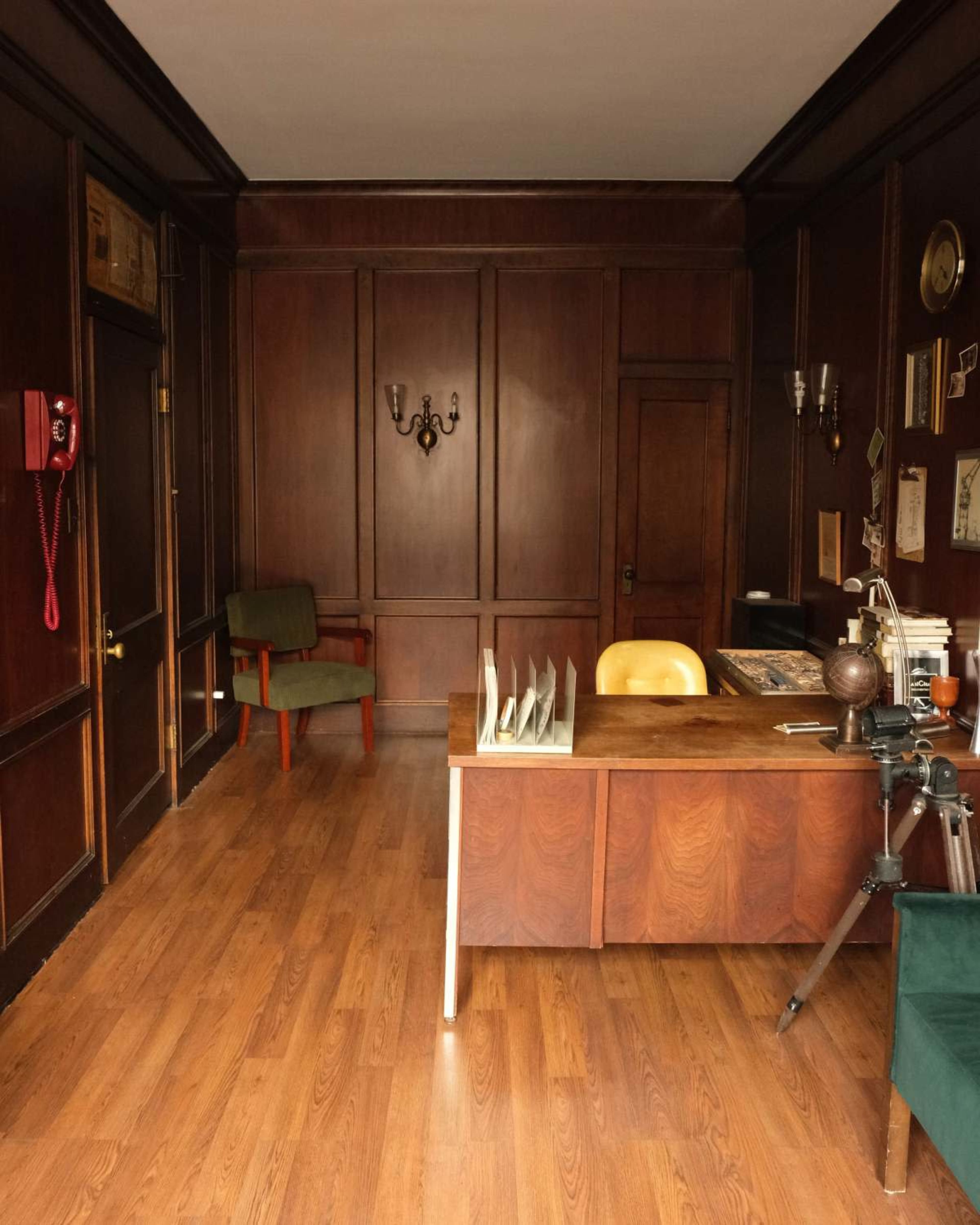 The image shows a wooden-paneled office with a desk, a green chair, and a vintage telephone mounted on the wall.