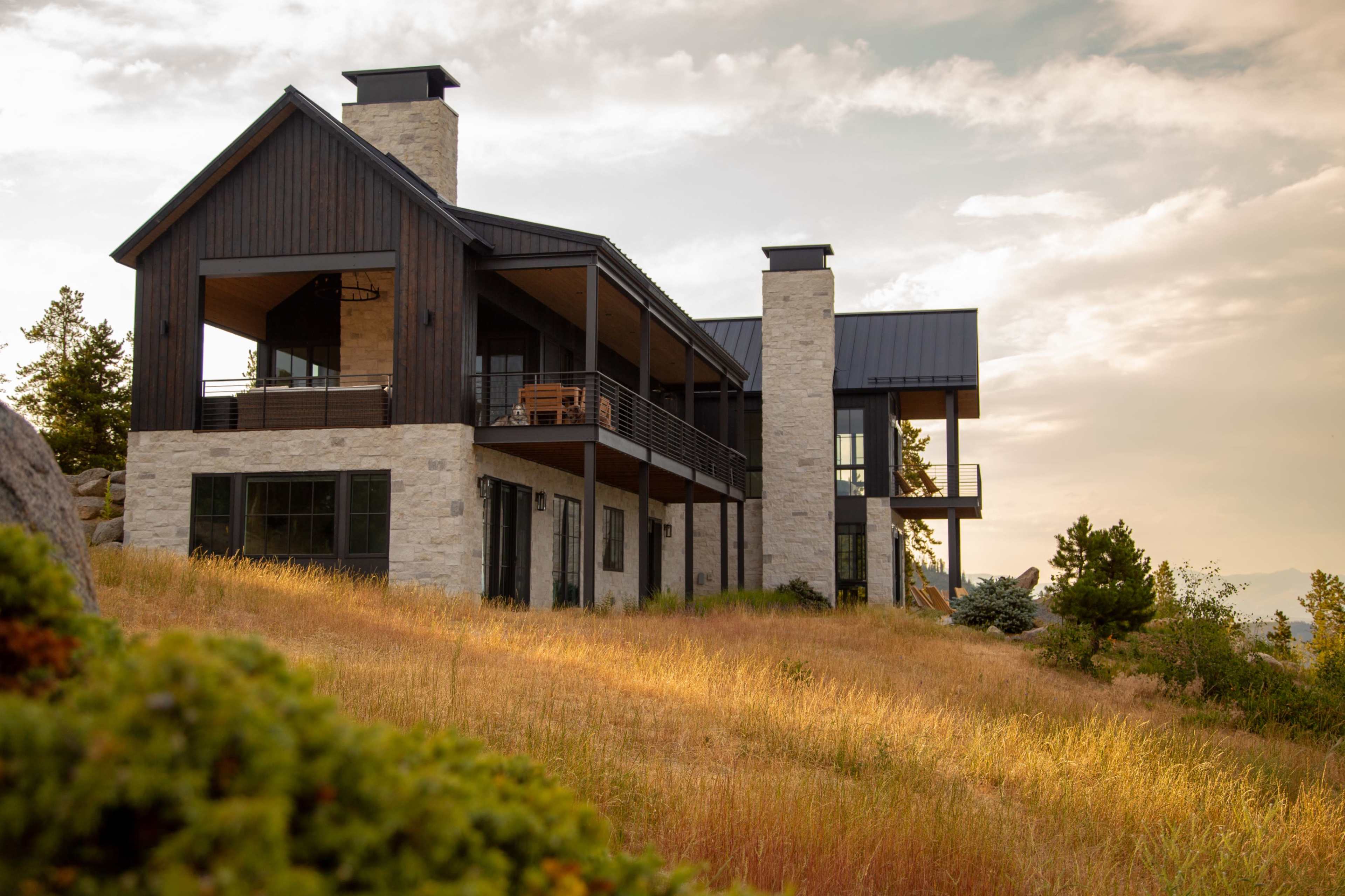 The image shows a modern house with a mix of stone and wood exteriors, situated on a grassy hill during sunset.