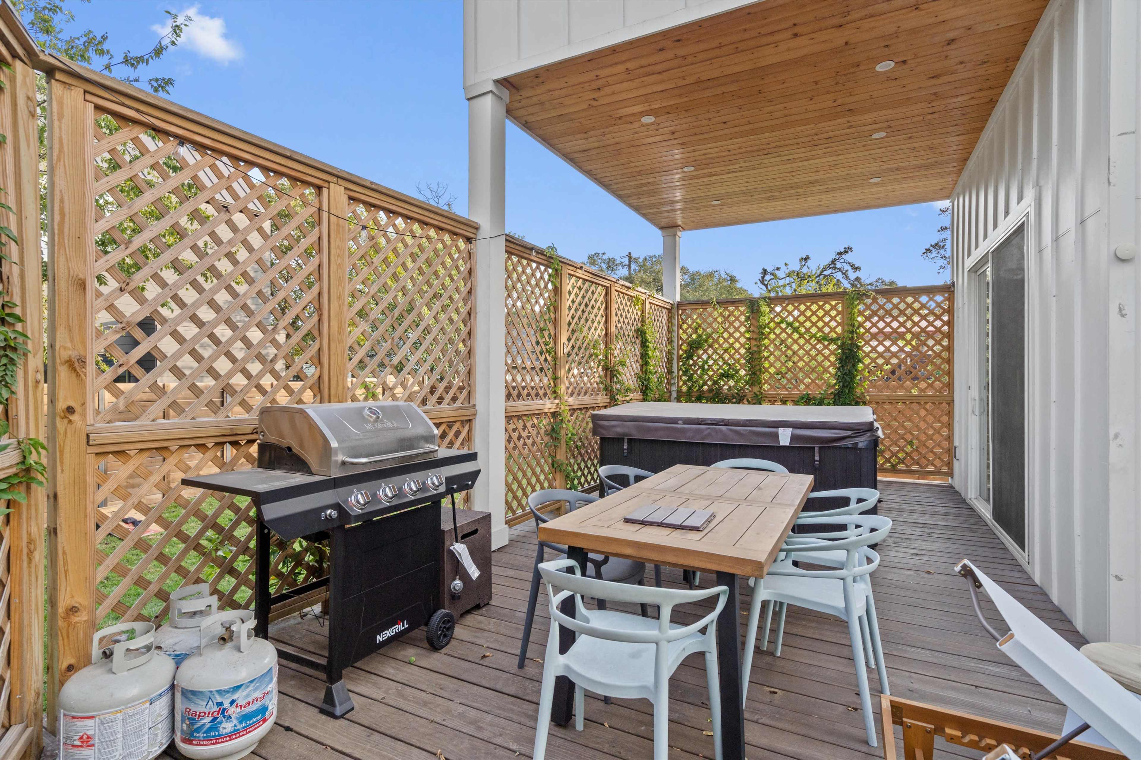 The image shows a wooden deck with a dining table, chairs, a gas grill, and a hot tub, surrounded by lattice fencing and greenery.